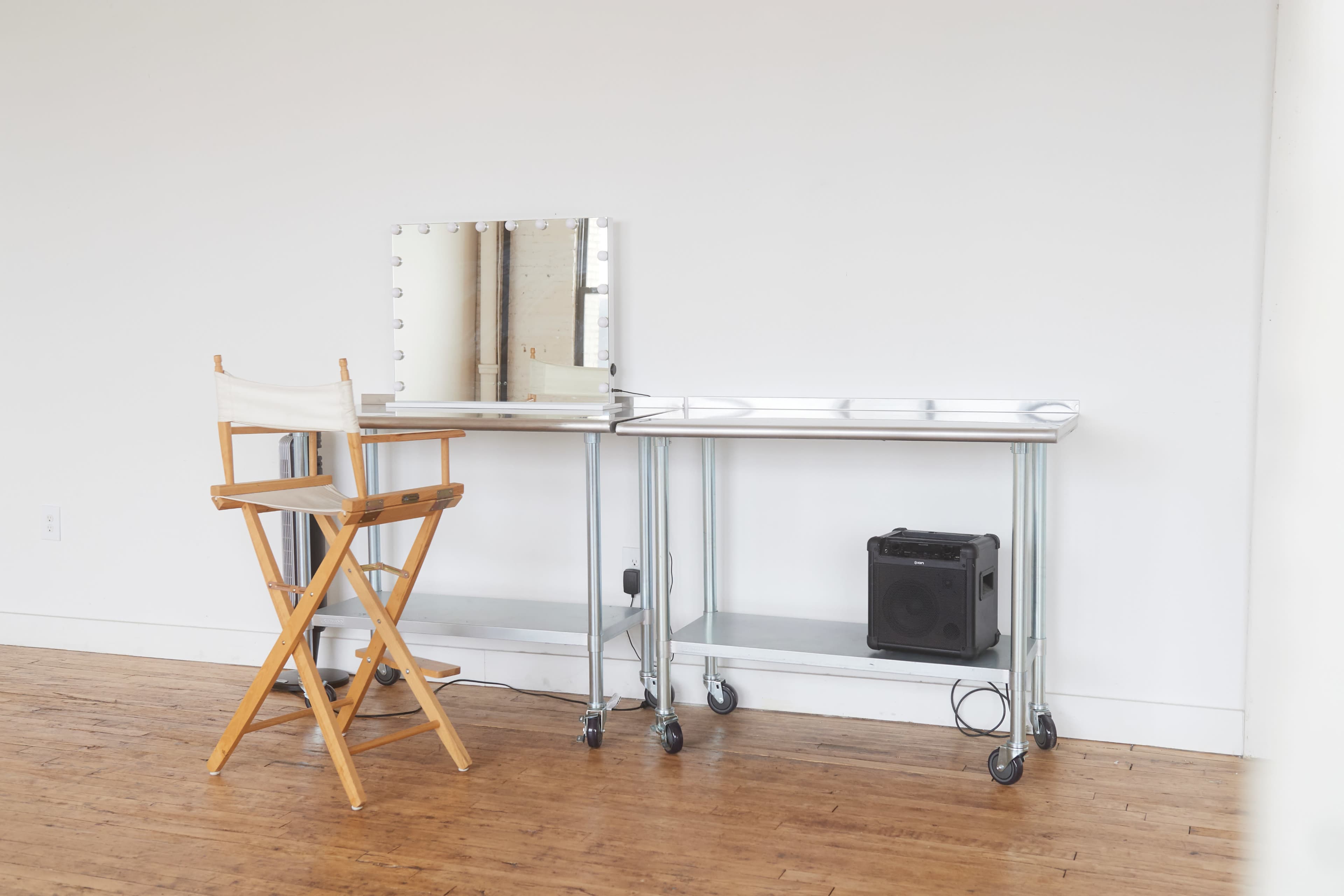 A makeup station with a mirror, a director's chair, and a small black speaker is set against a plain white wall.