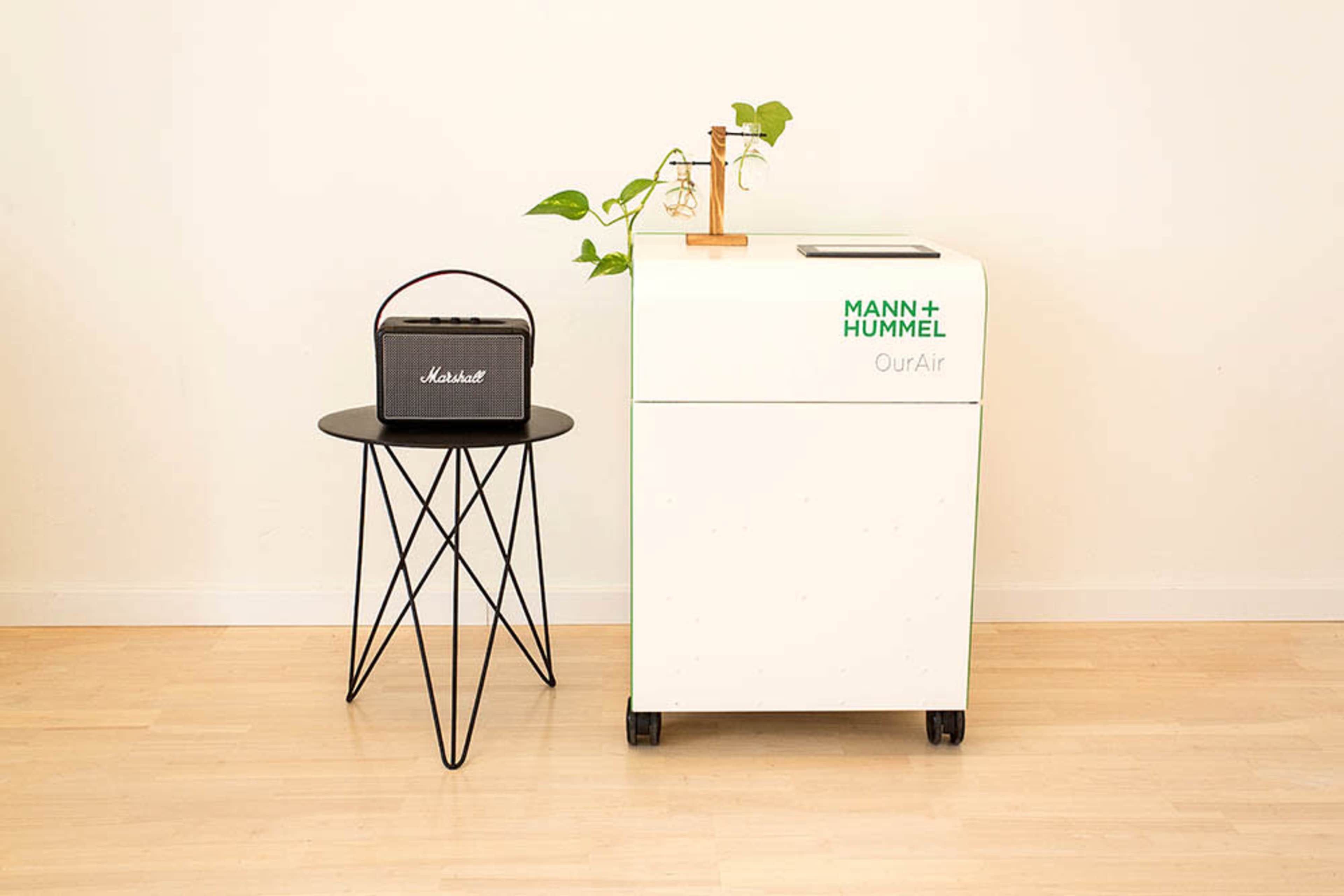 A modern air purifier on wheels next to a small black speaker on a round, wire-framed table, with a plant in a pot on the table.