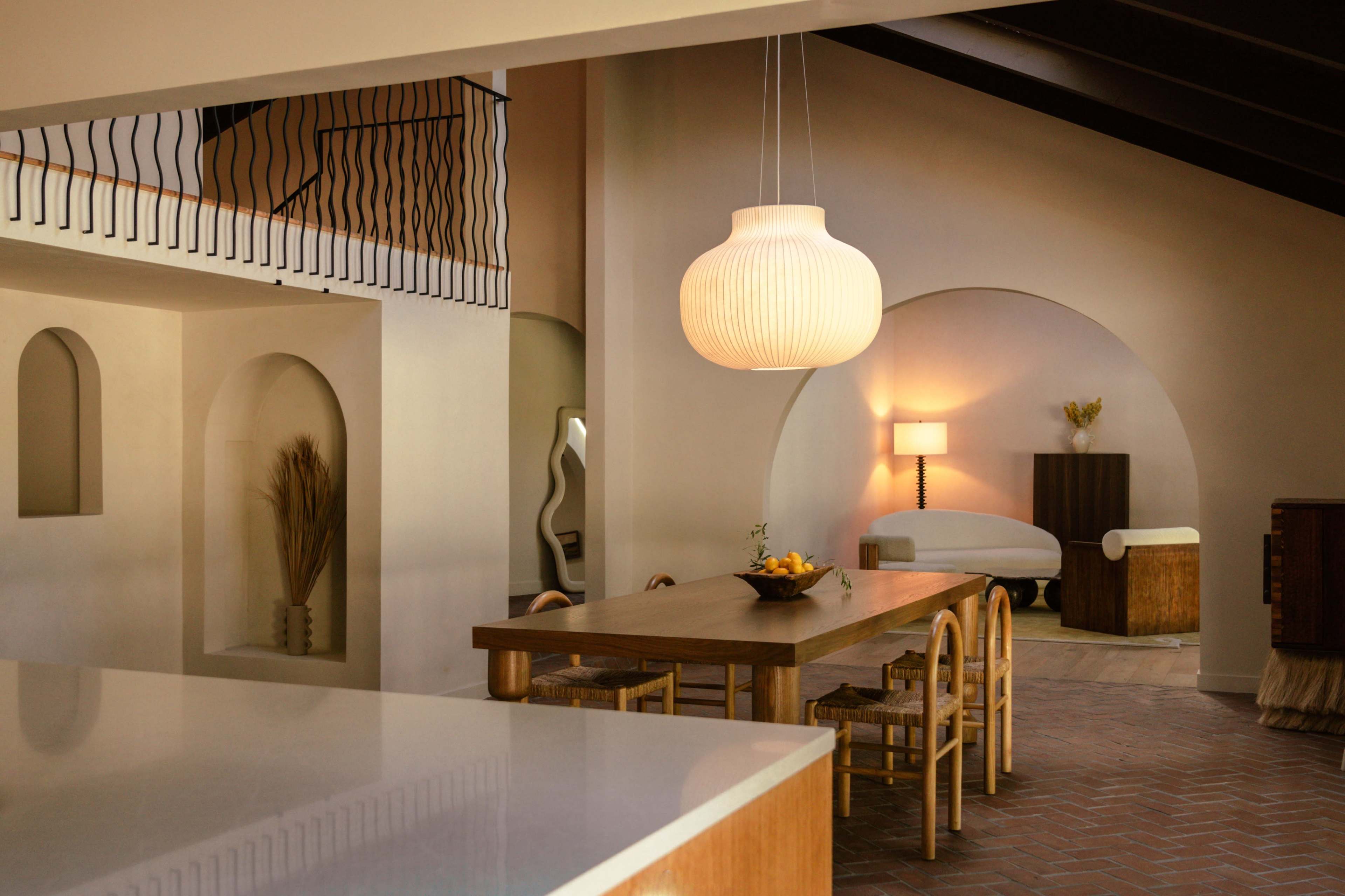 The image shows a modern interior space with a dining area featuring a large wooden table, woven chairs, and a prominent pendant light, alongside curved architectural elements and soft furnishings.