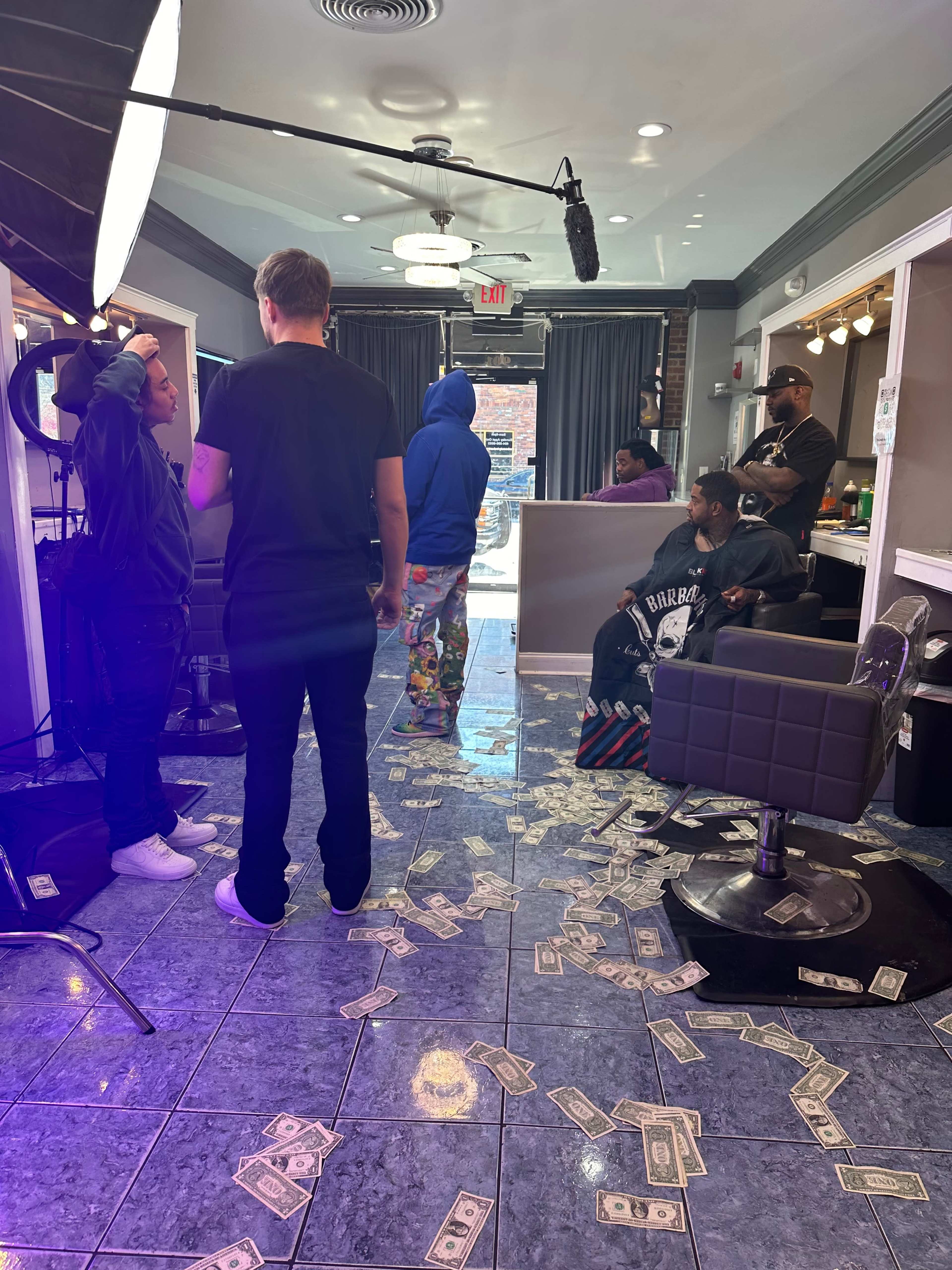 A barbershop interior is decorated with scattered bills on the floor as customers and stylists engage in conversation.