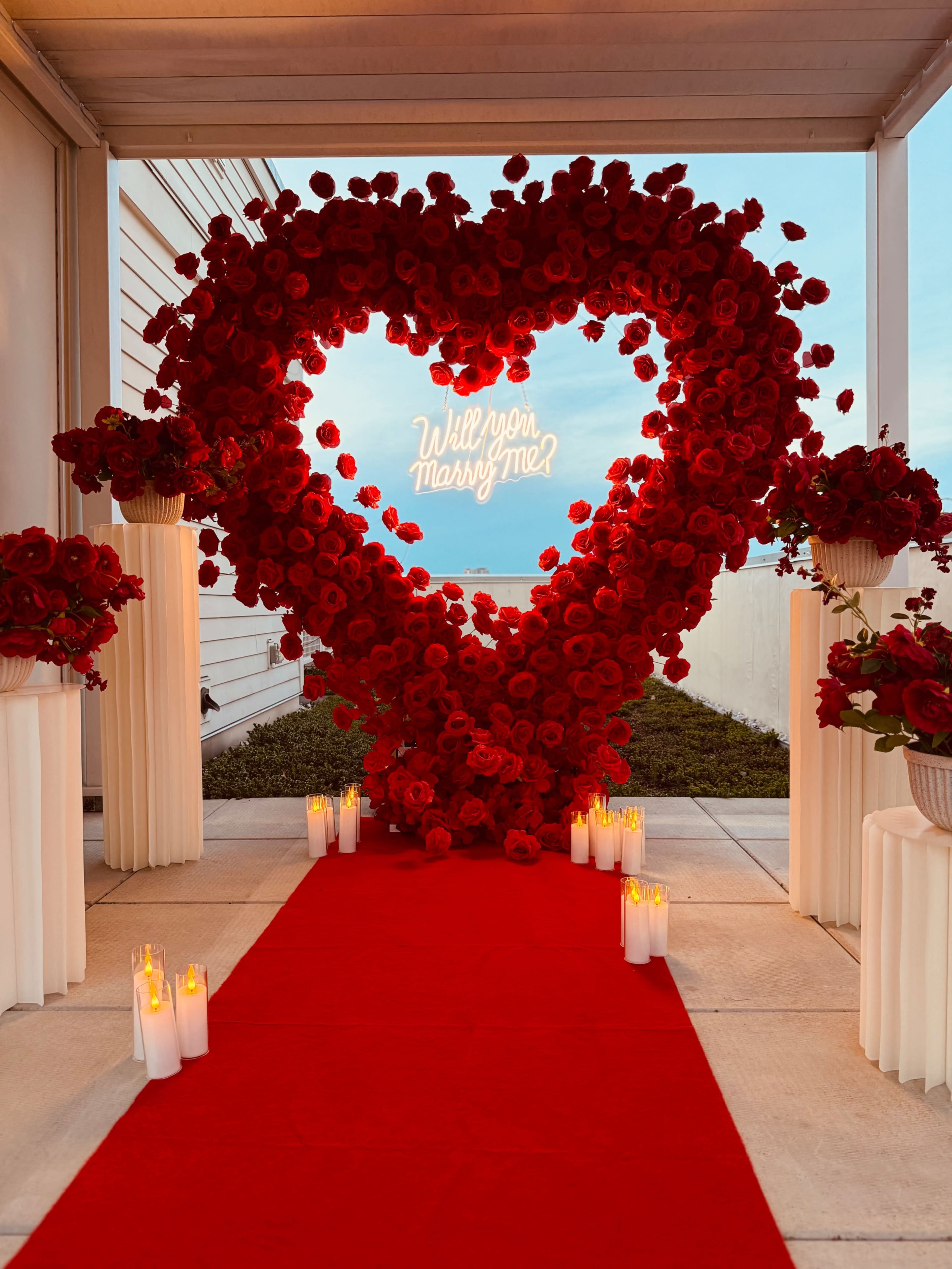 A heart-shaped floral display made of red roses is illuminated by a neon sign saying "Will you Marry Me?" in front of a pathway lined with candles and a red carpet.