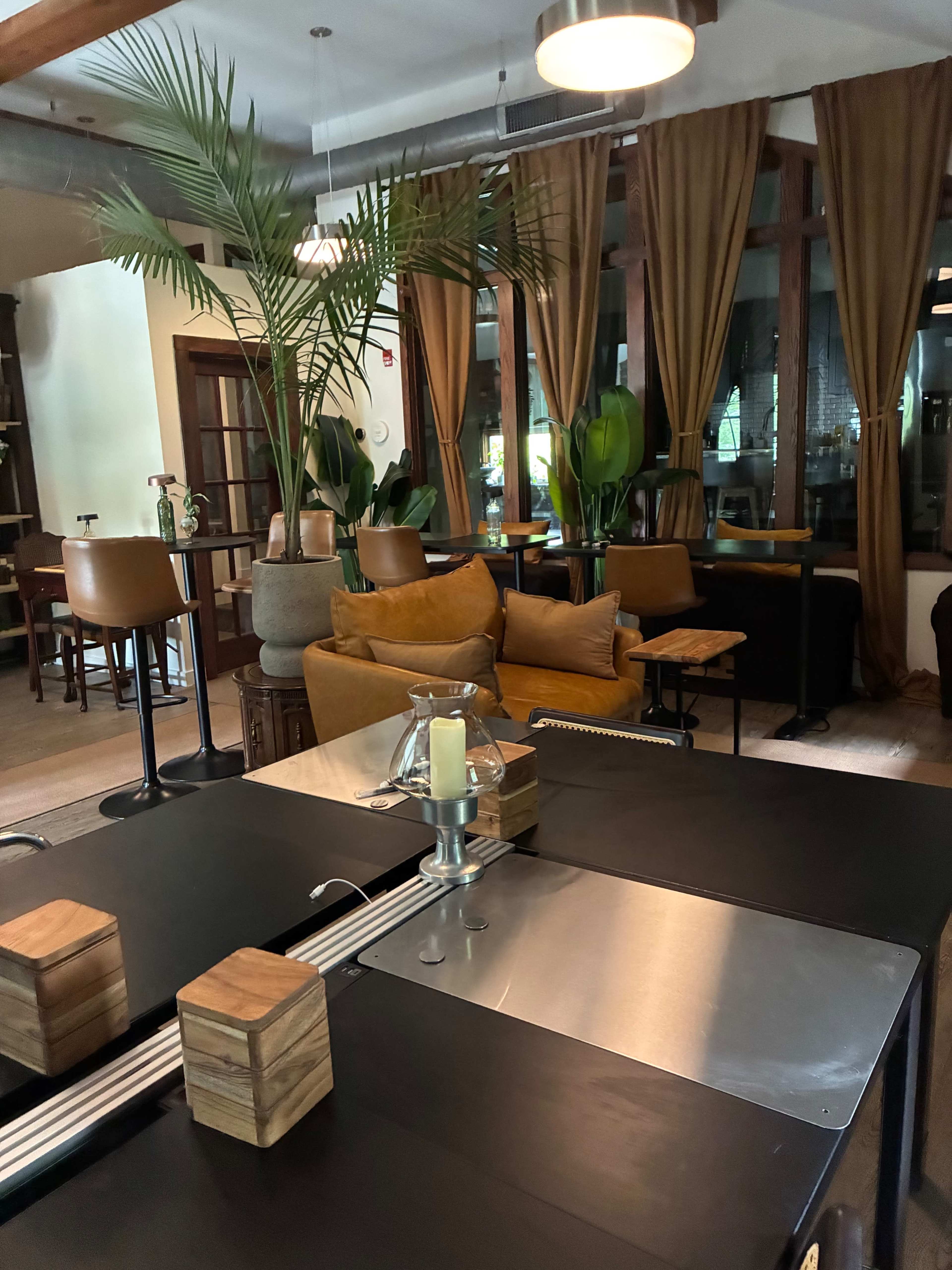 The image shows an interior space featuring dark tables, wooden accents, plants, and seating areas with large windows dressed in brown curtains.