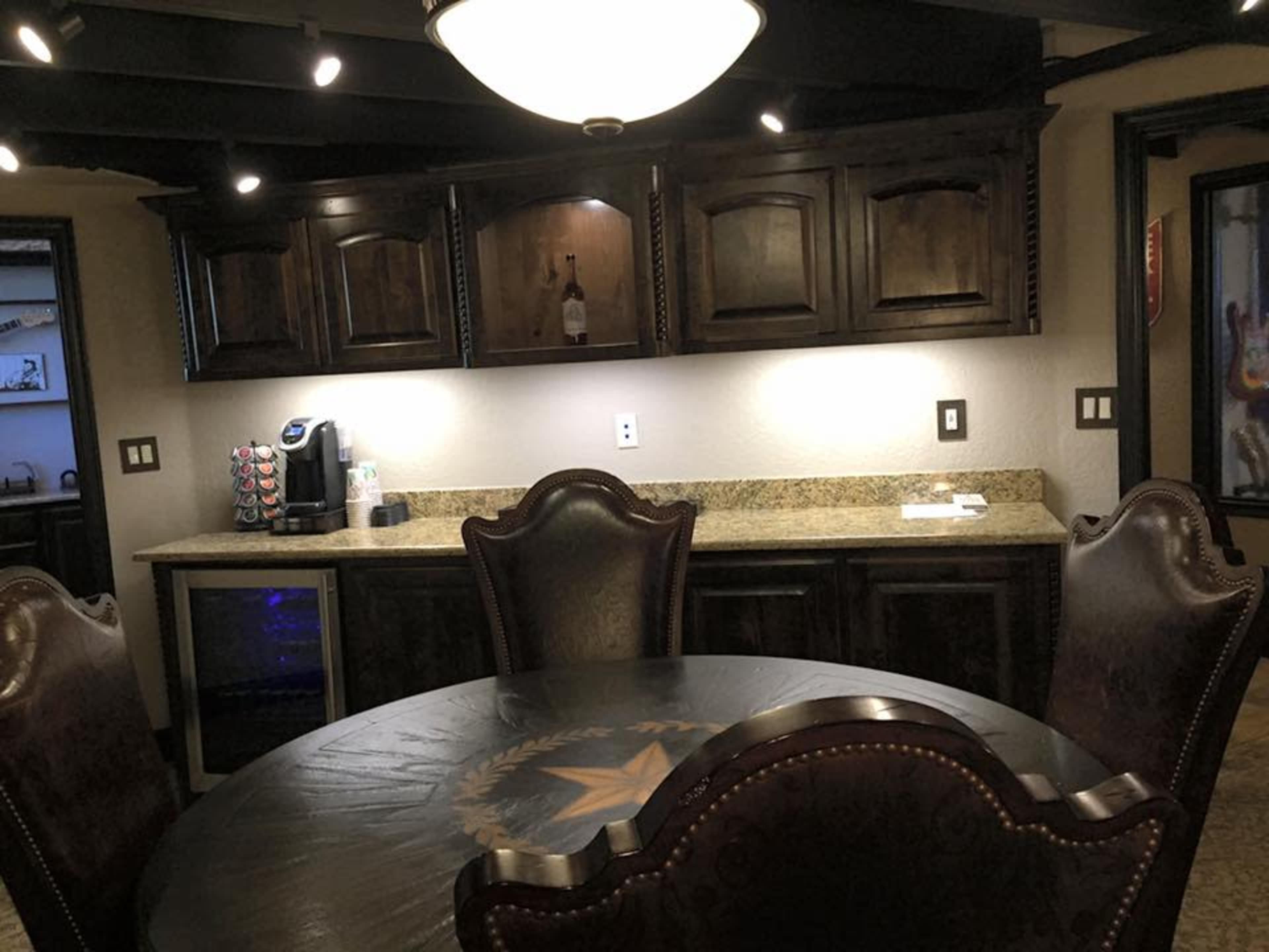 The image shows a dark wooden kitchen with a round table, four ornate chairs, and a countertop featuring a coffee maker and a small refrigerator.