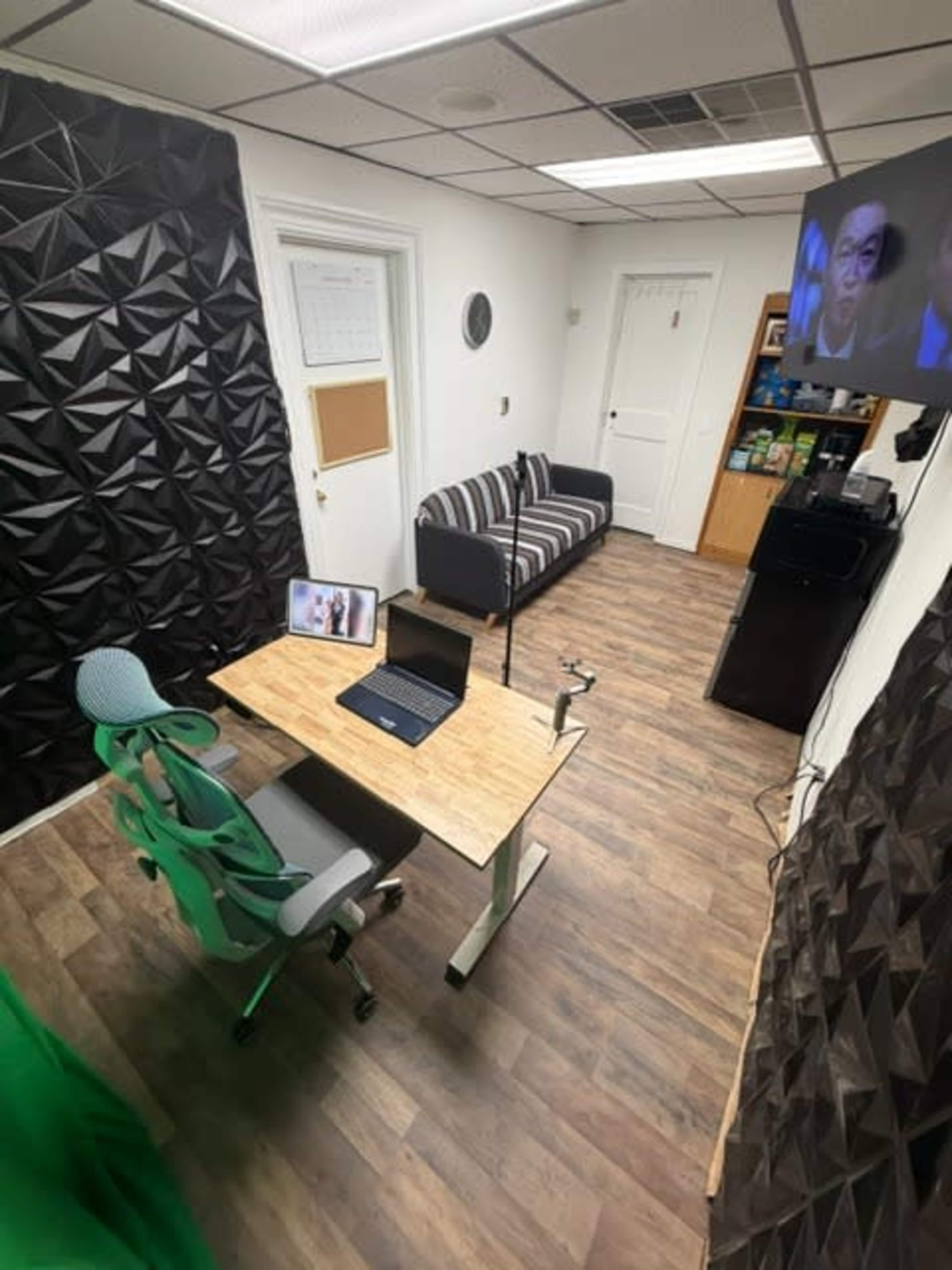The image shows a small office space featuring a wooden desk with a laptop, an ergonomic chair, a couch, and a television mounted on the wall.