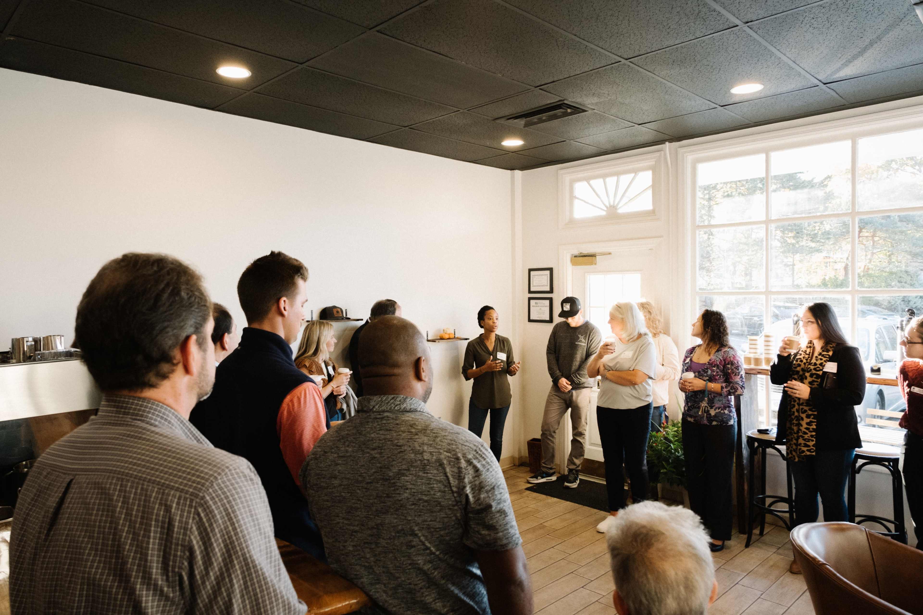 A group of people stands in a brightly lit room, engaging in conversation near a large window.
