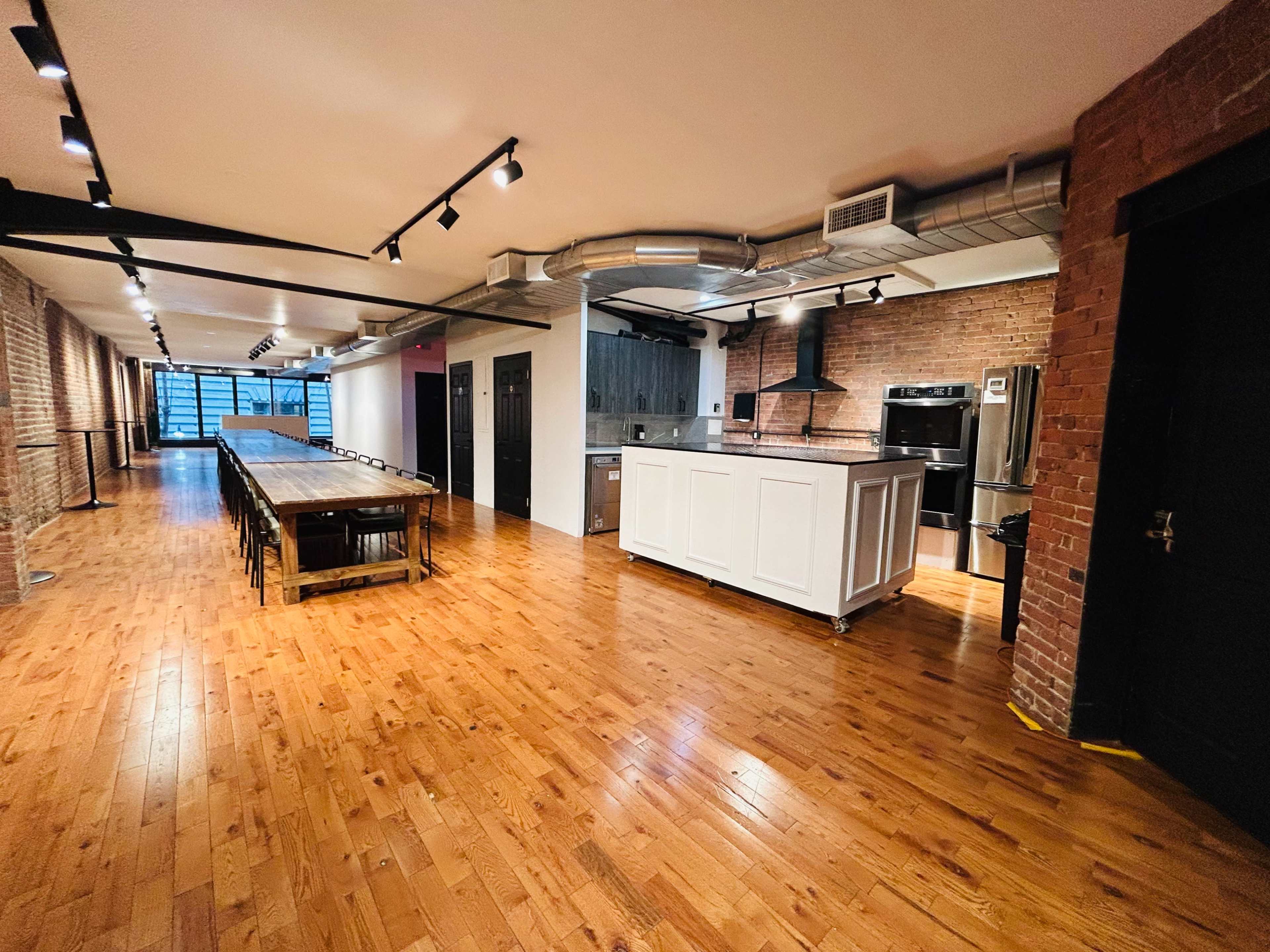 The image features a spacious kitchen and dining area with wooden flooring, exposed brick walls, and a large communal table positioned alongside a modern kitchen setup.