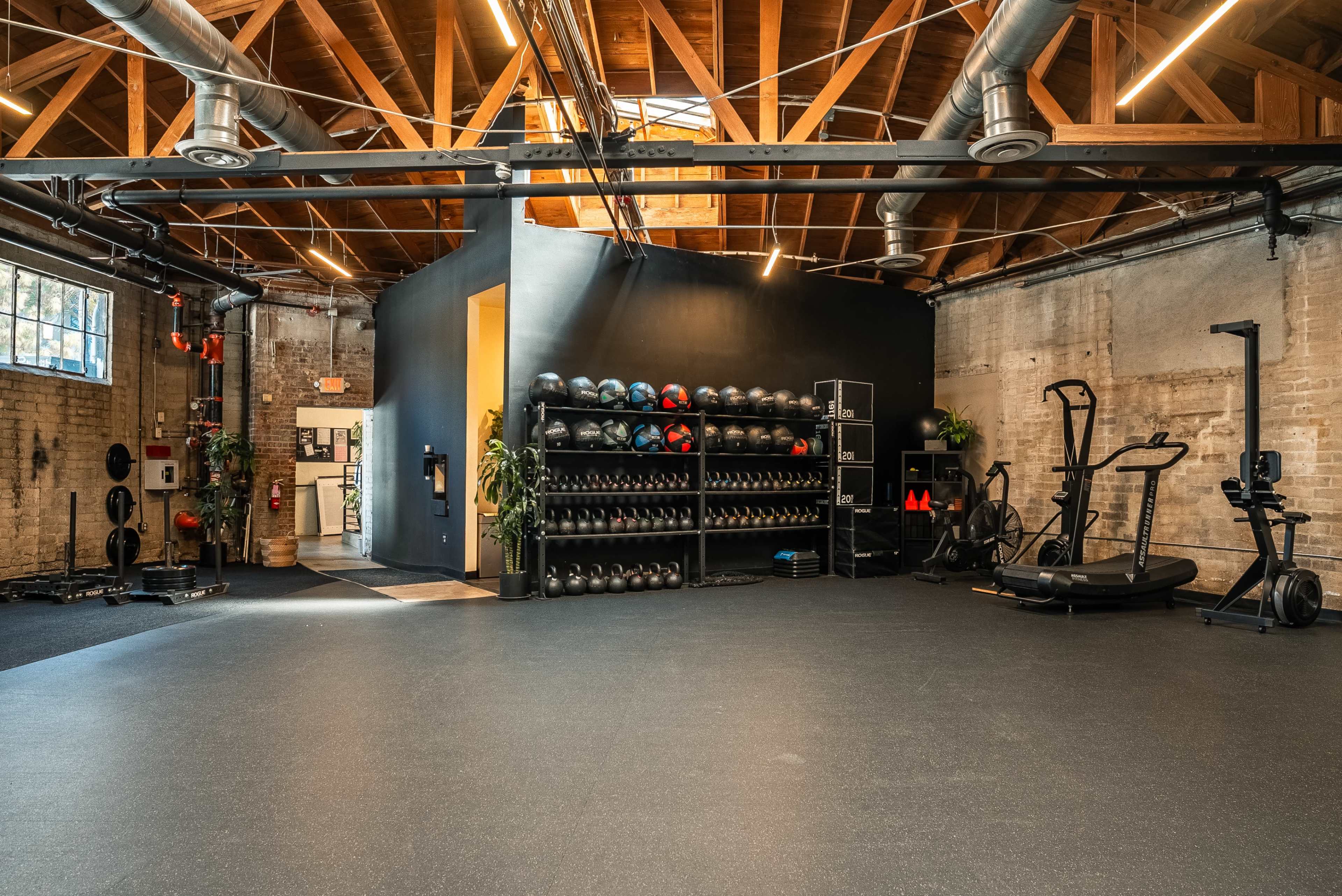 The image shows a spacious gym with wooden beams, black walls, and various exercise equipment, including weights and cardio machines, organized along the walls.
