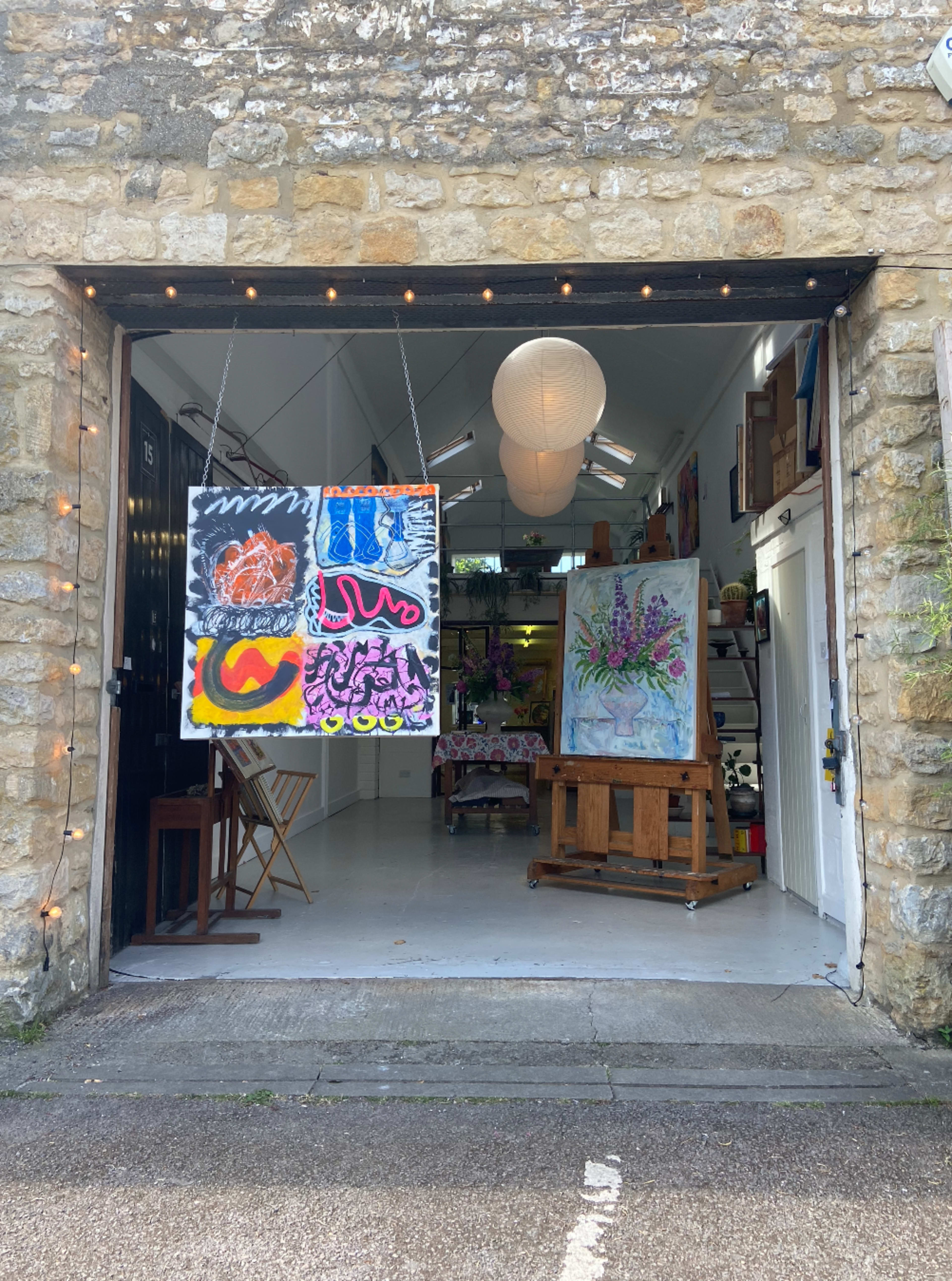 The image shows an art studio with open doors displaying colorful artworks and an easel holding a floral painting.