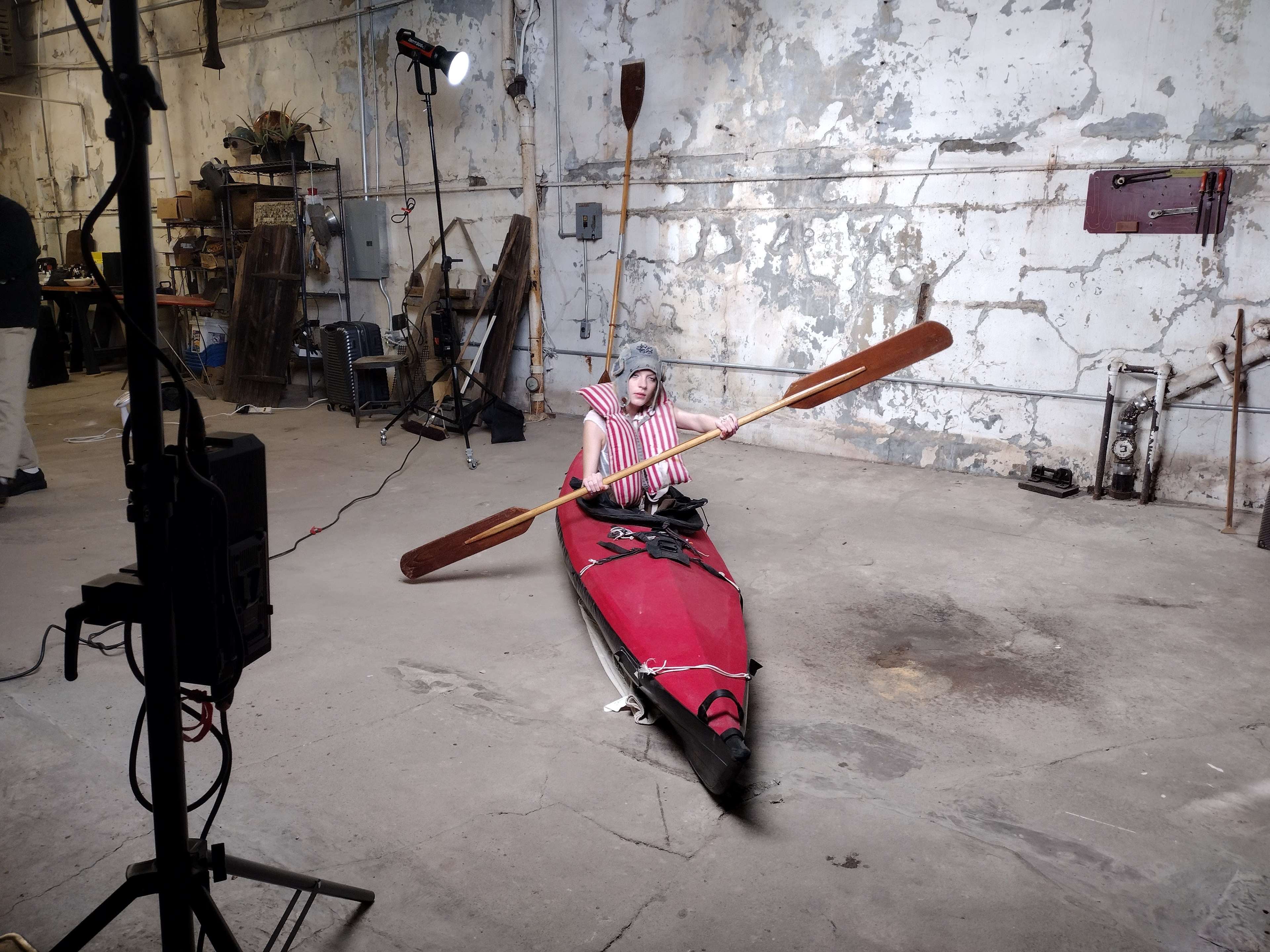 A person in a striped outfit is sitting in a red kayak, holding paddles, inside a warehouse with exposed walls and various tools in the background.