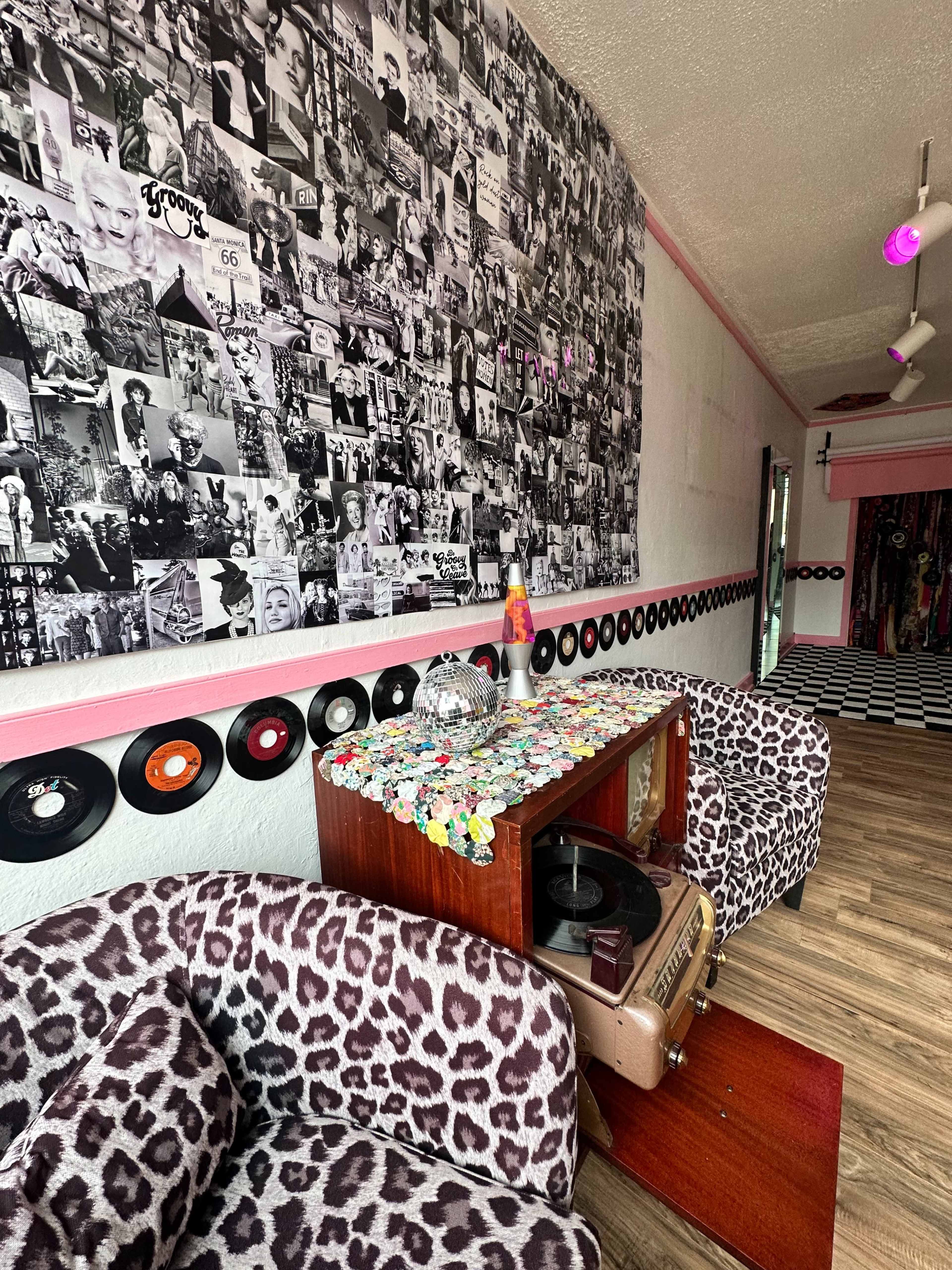The room features a wall covered in black and white photos, with a vintage record player and patterned chairs in front, surrounded by colorful vinyl records.