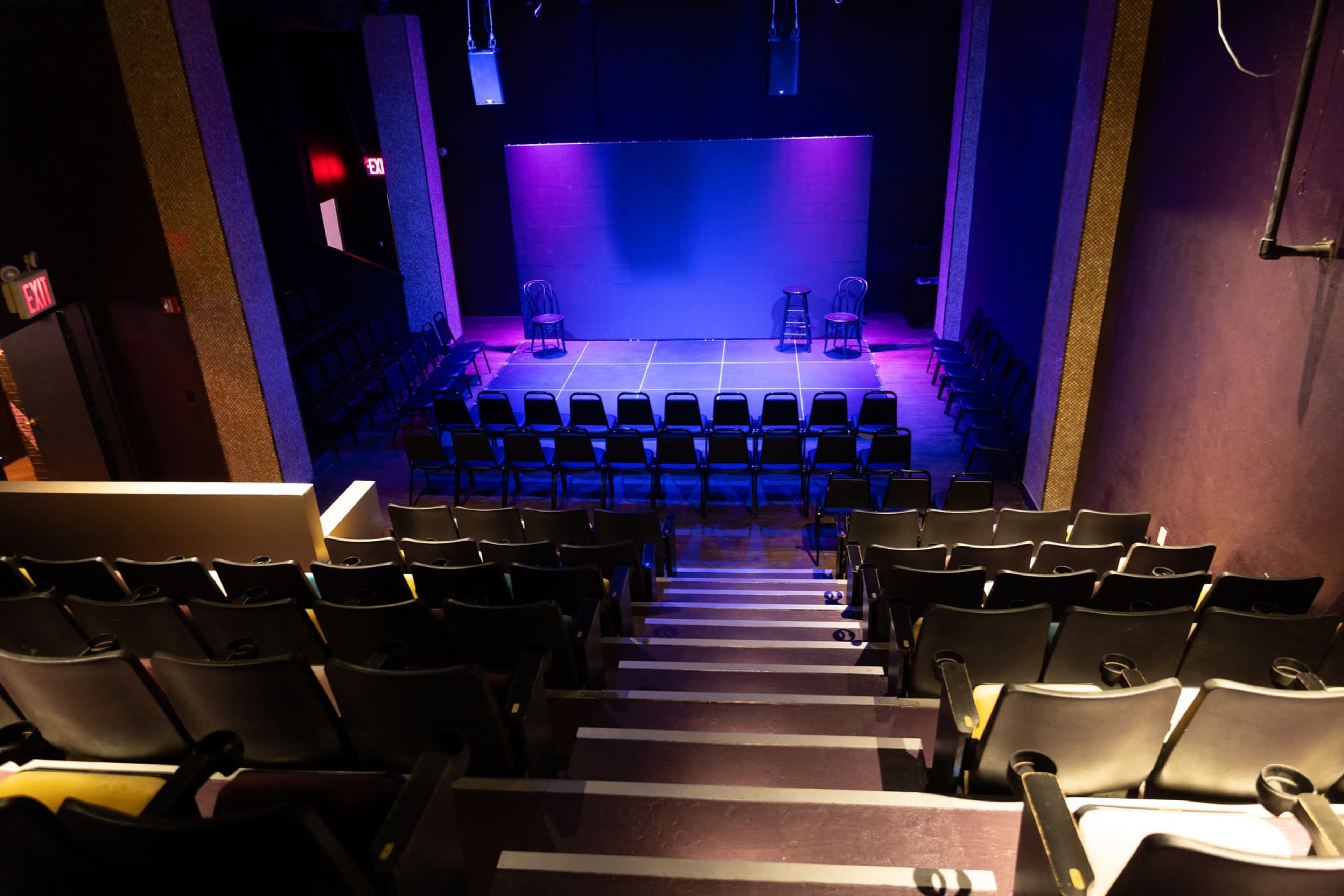 An empty theater with rows of black chairs facing a small stage illuminated with purple lighting.