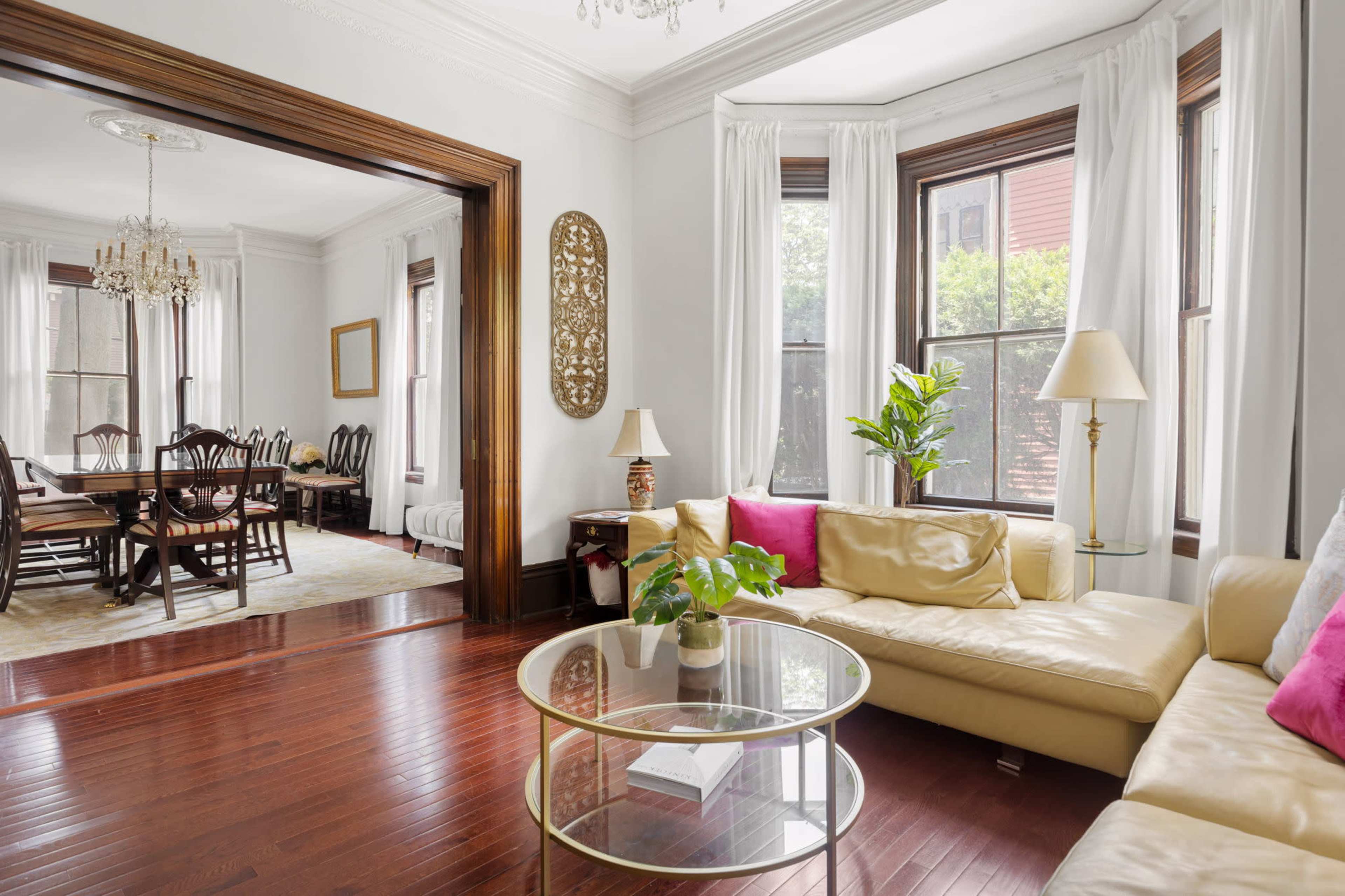 A living room features a beige sectional sofa, a round glass coffee table with a plant, and large windows allowing natural light, with a dining area visible in the background.