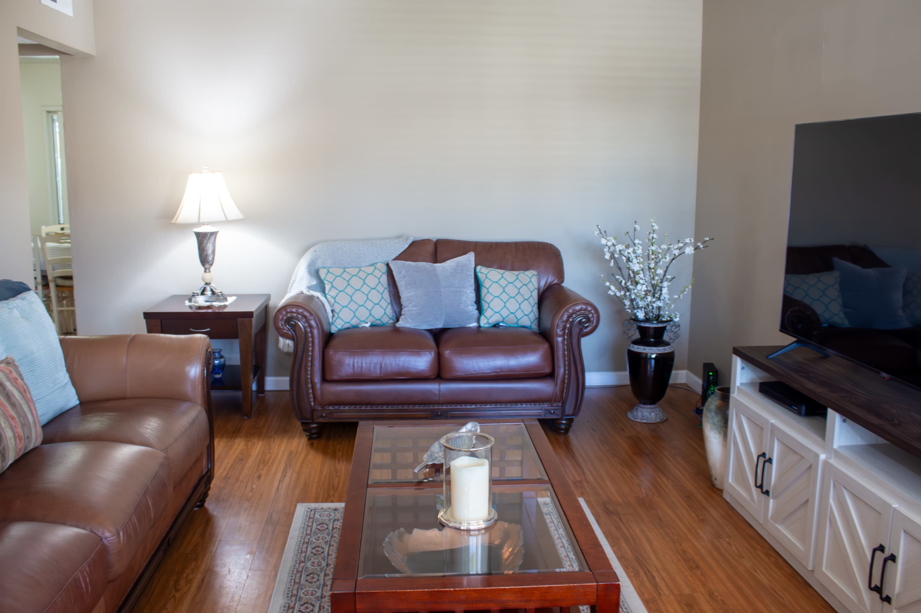 The living room features a brown leather sofa, a wooden coffee table with a candle, a side table with a lamp, and a television stand with a TV.