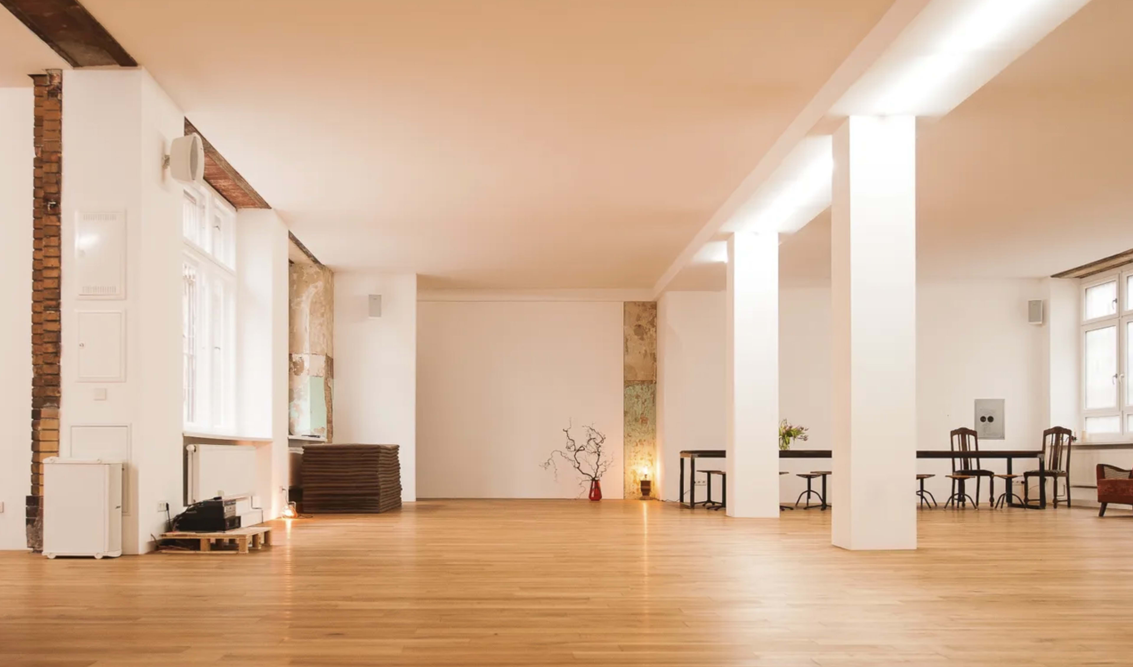 A spacious, minimalist indoor area with wooden flooring, white walls, and a few tables and chairs arranged in a simple layout.