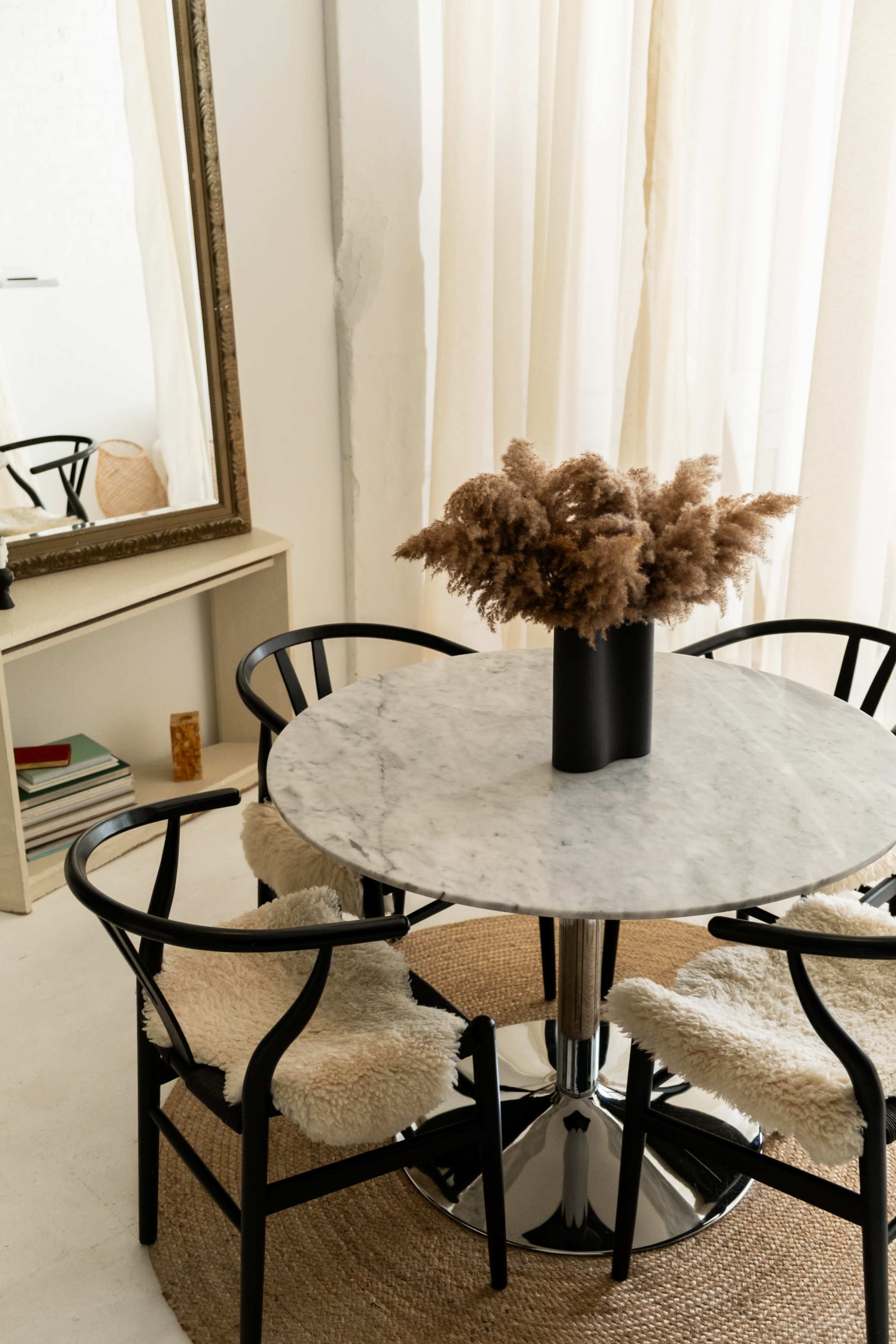A round marble table with four black chairs surrounds a vase of dried plants, positioned next to a large mirror and light-colored curtains.