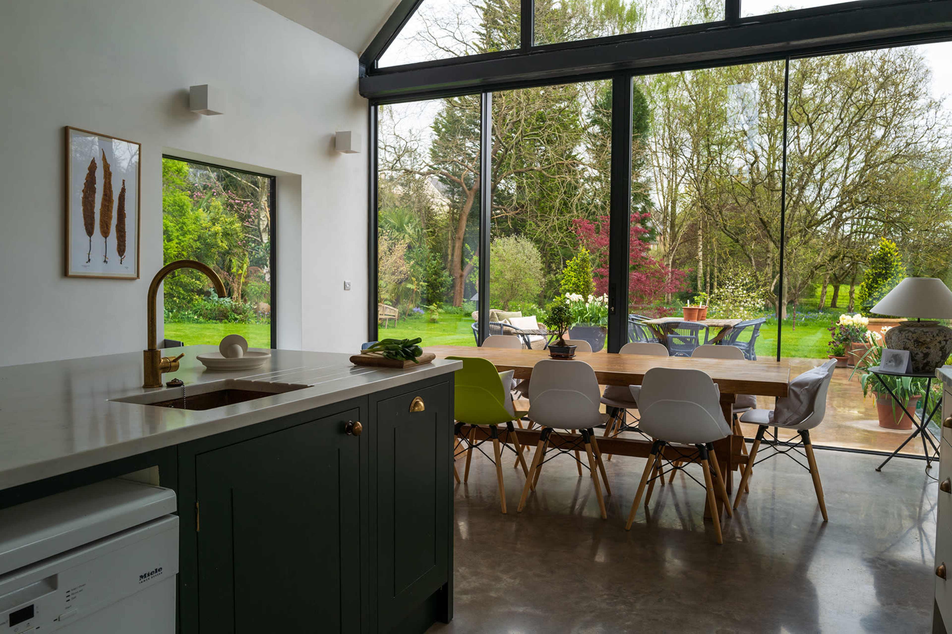 A modern kitchen with a green island and a dining area overlooks a lush garden through large glass windows.