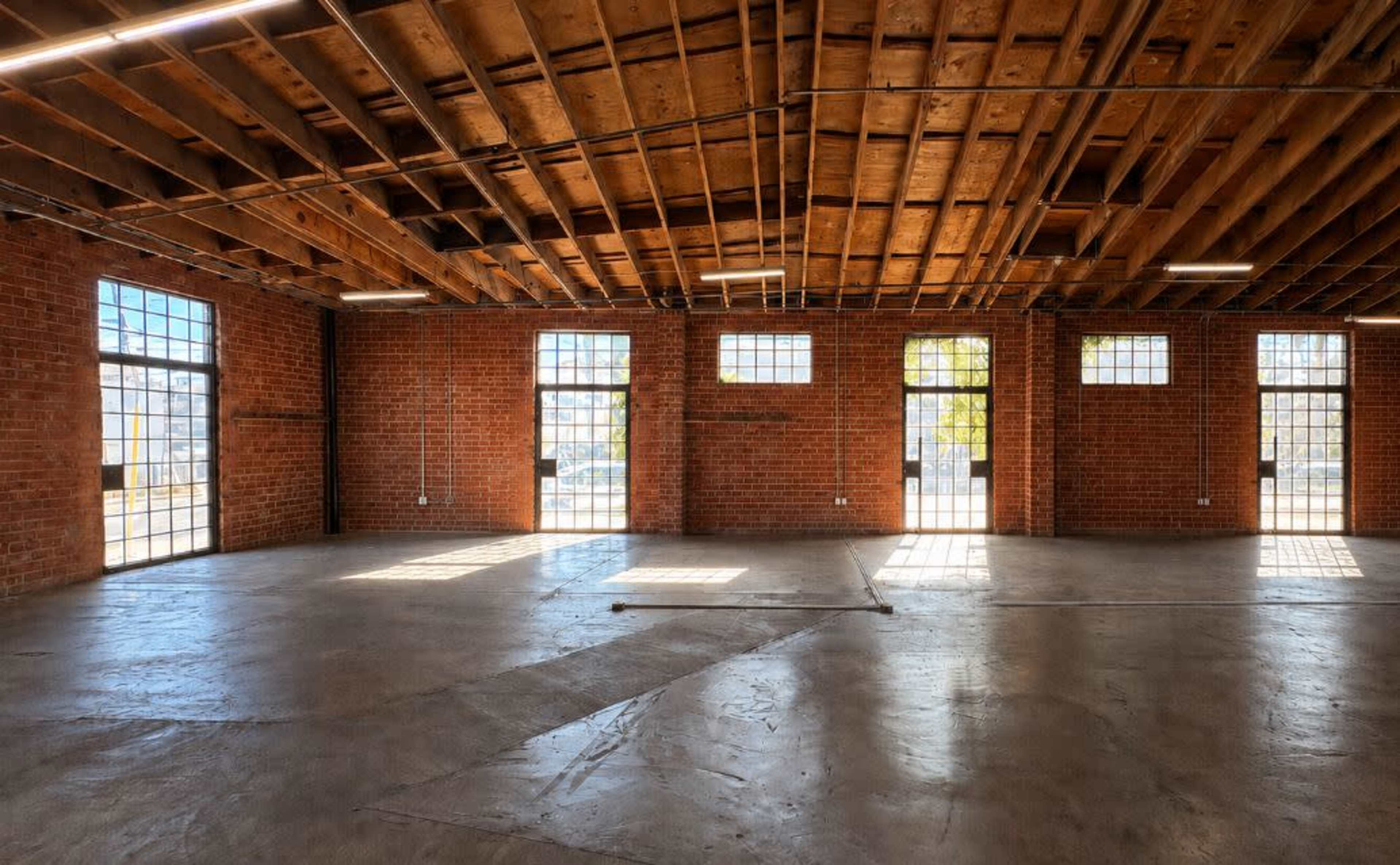 The image shows an empty industrial space with exposed brick walls, a high wooden ceiling, and large windows allowing natural light to enter.