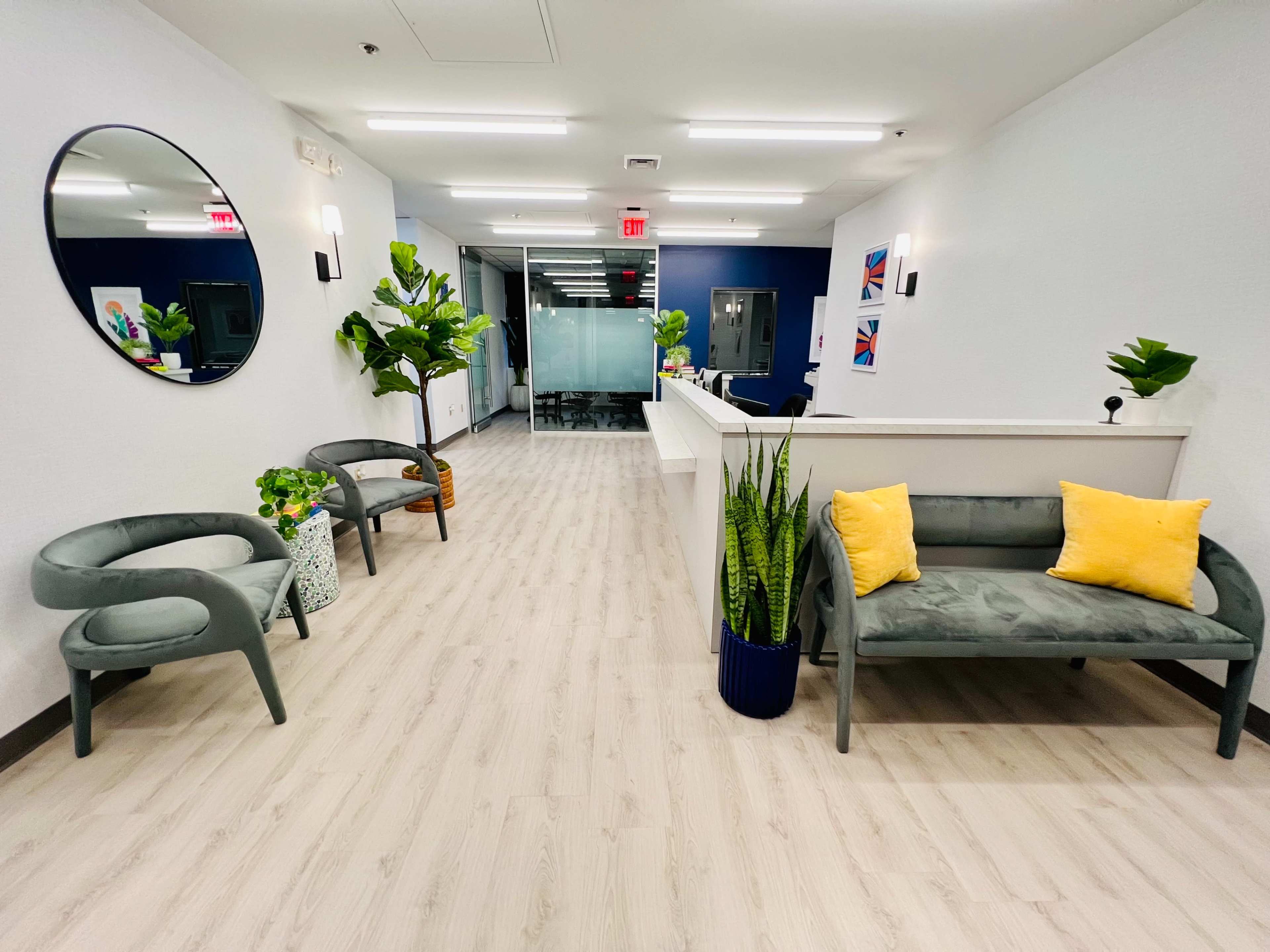 A modern office lobby with two green chairs, a gray sofa with yellow cushions, potted plants, and a reception desk at the far end.