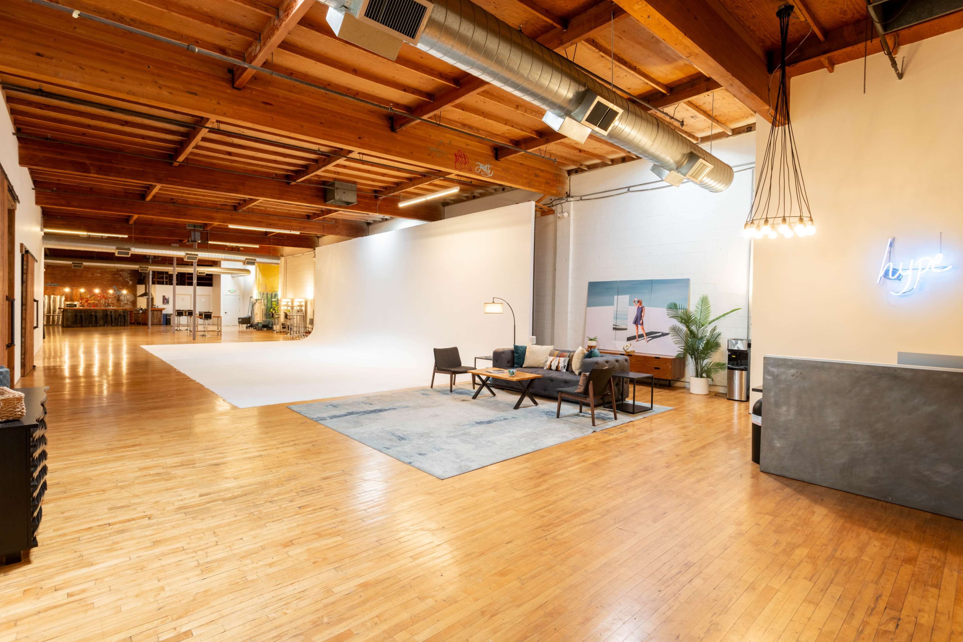 The image shows a spacious, well-lit interior with wooden beams, a large white backdrop area, and modern furniture arranged in a lounge setting.
