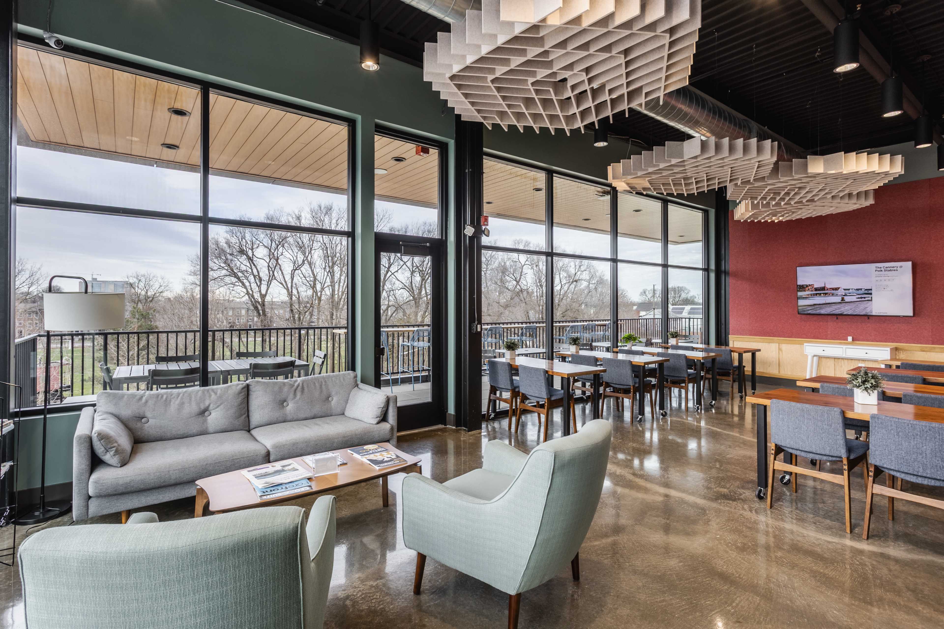 The image shows a modern lounge area with large windows, stylish seating, and a dining space that overlooks a landscaped outdoor area.