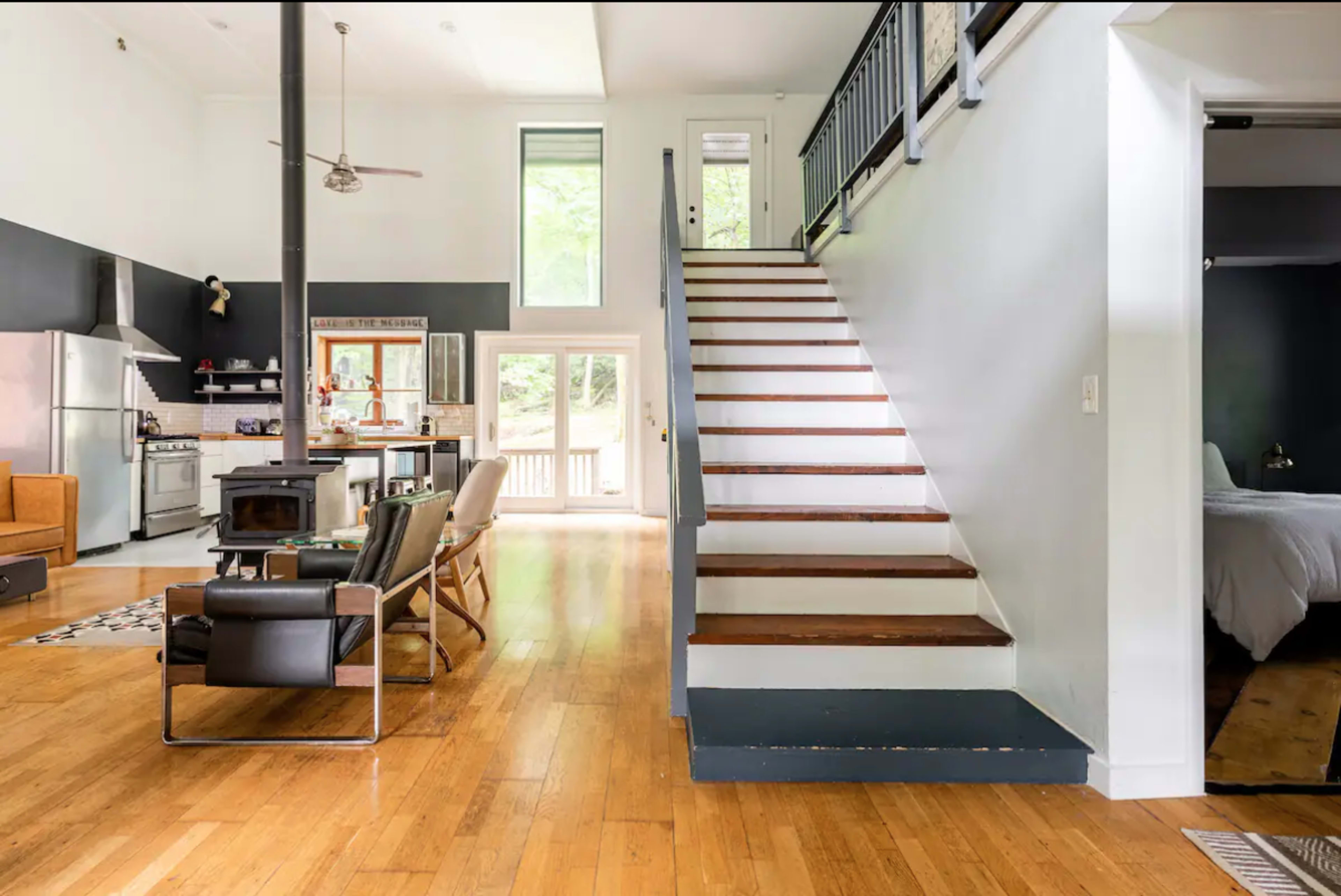 The image shows an open-concept living space featuring a kitchen, dining area, and a staircase leading to an upper level, with large windows allowing natural light to enter.