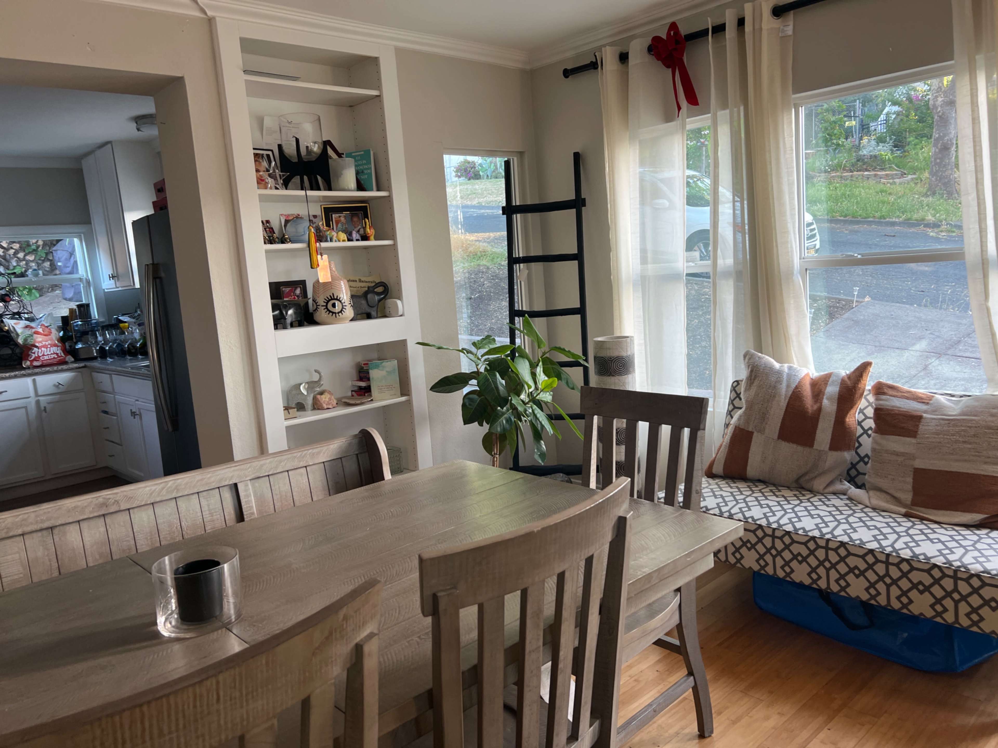 A cozy dining area with a wooden table, chairs, and a window seat adorned with cushions, alongside a small plant and shelves displaying various items.