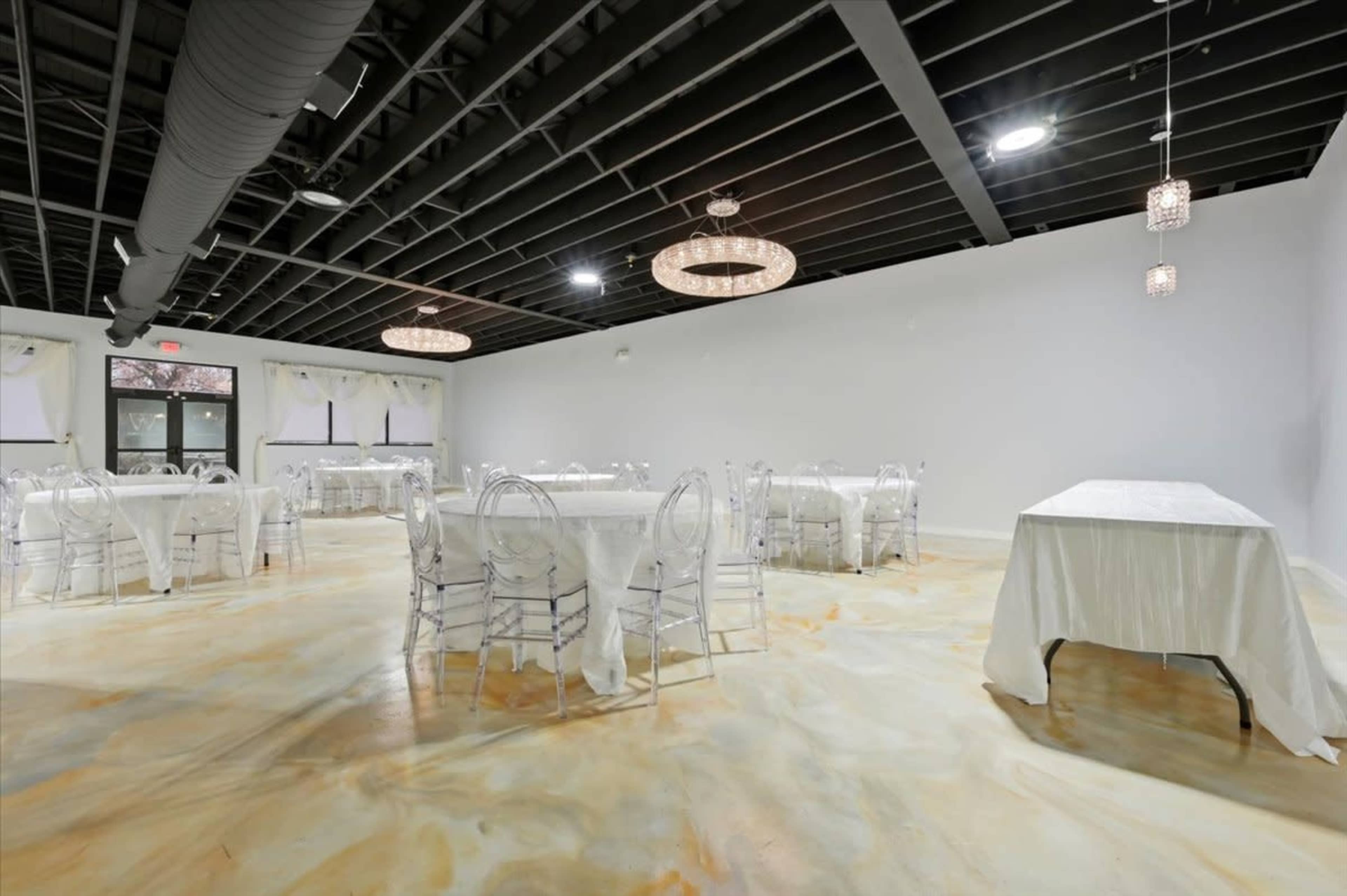 The image shows an empty event space with several round tables covered in white tablecloths and modern chandeliers hanging from the ceiling.