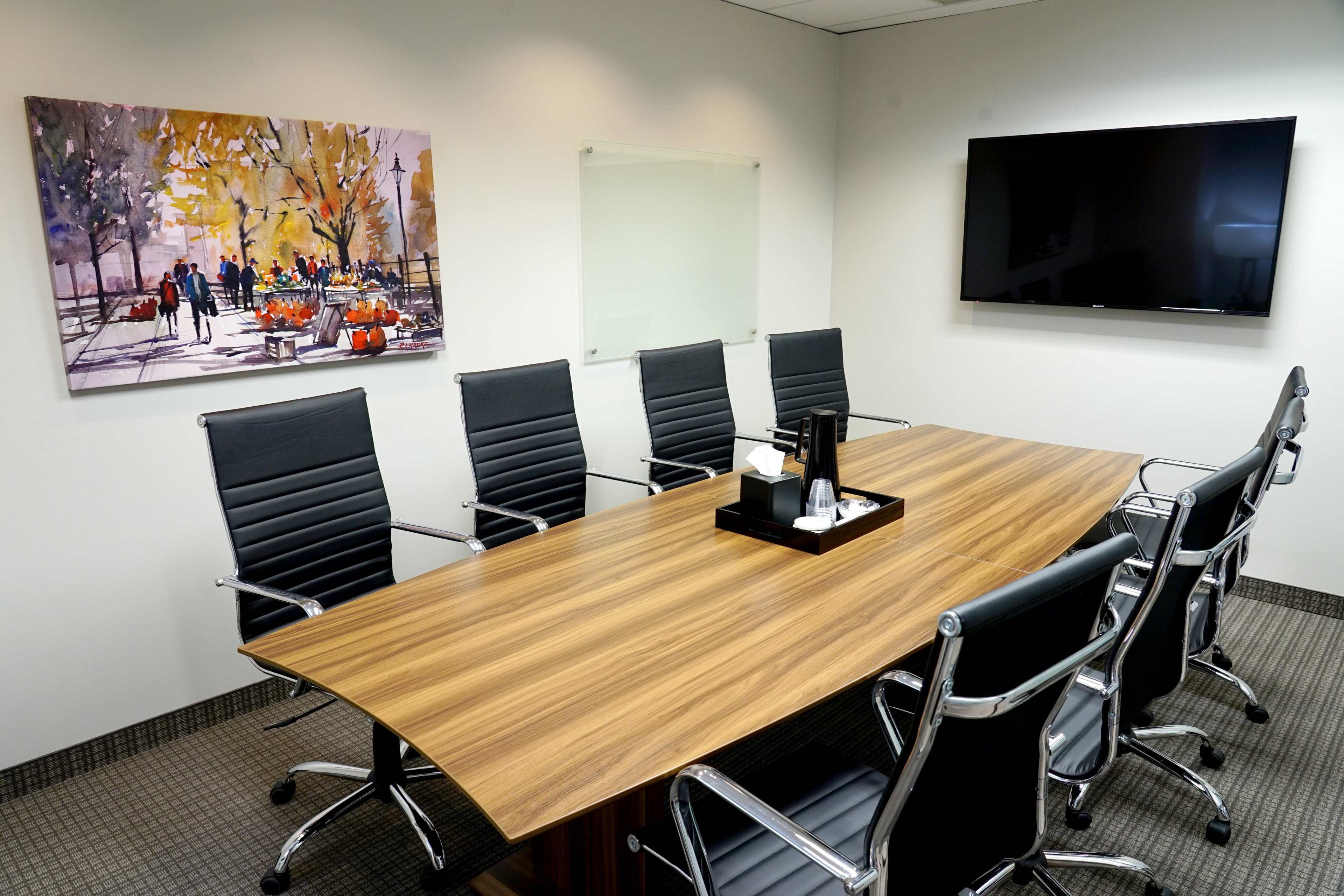 A modern conference room features a long wooden table surrounded by six black chairs, a wall-mounted television, and a colorful artwork depicting a park scene.