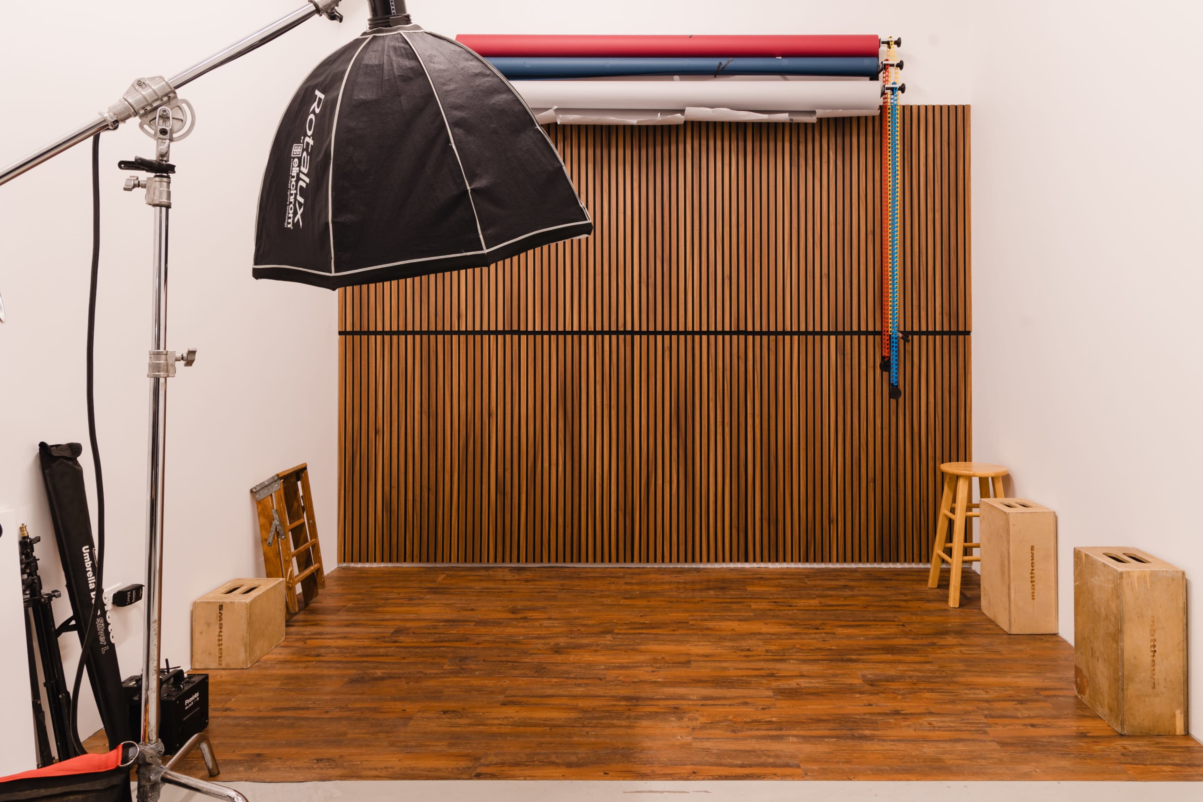 The image shows a photography studio with a wooden wall, lighting equipment, a stool, and props on the floor.