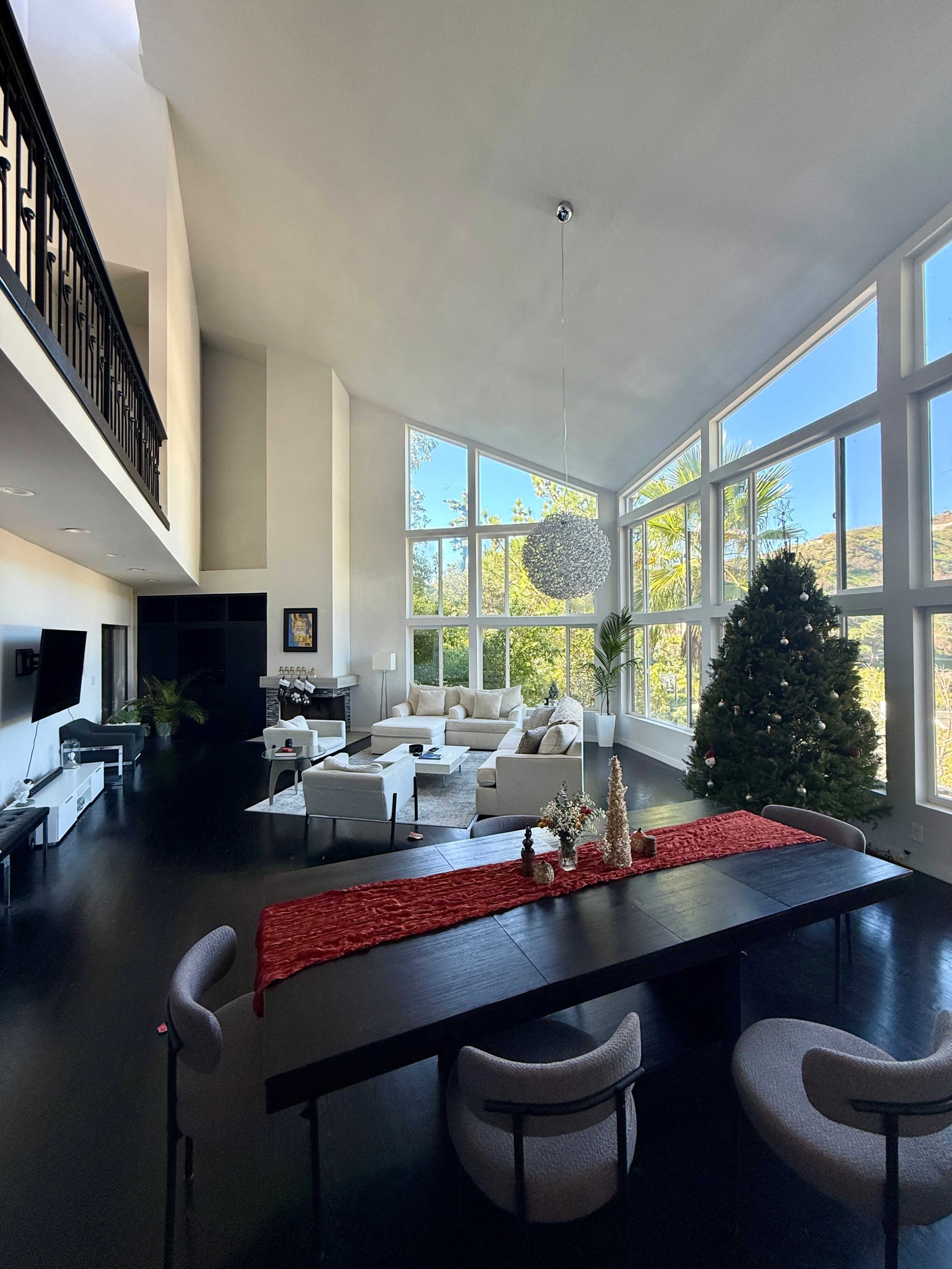 A spacious living area features large windows, a dining table with a red table runner, modern furniture, and a Christmas tree in the corner.
