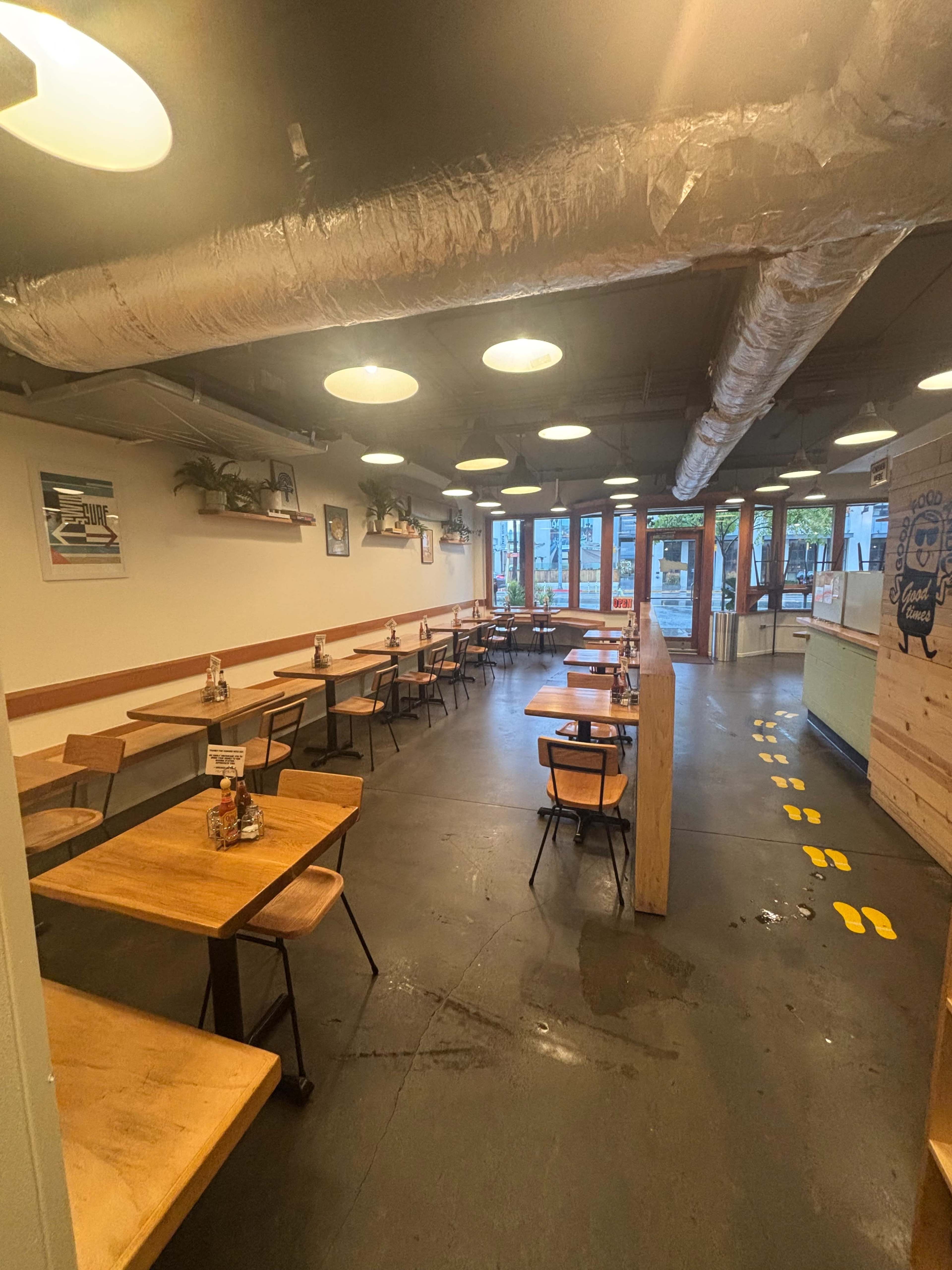 The image shows a spacious cafe interior with wooden tables and chairs arranged along the walls, and a row of lights hanging from the ceiling.