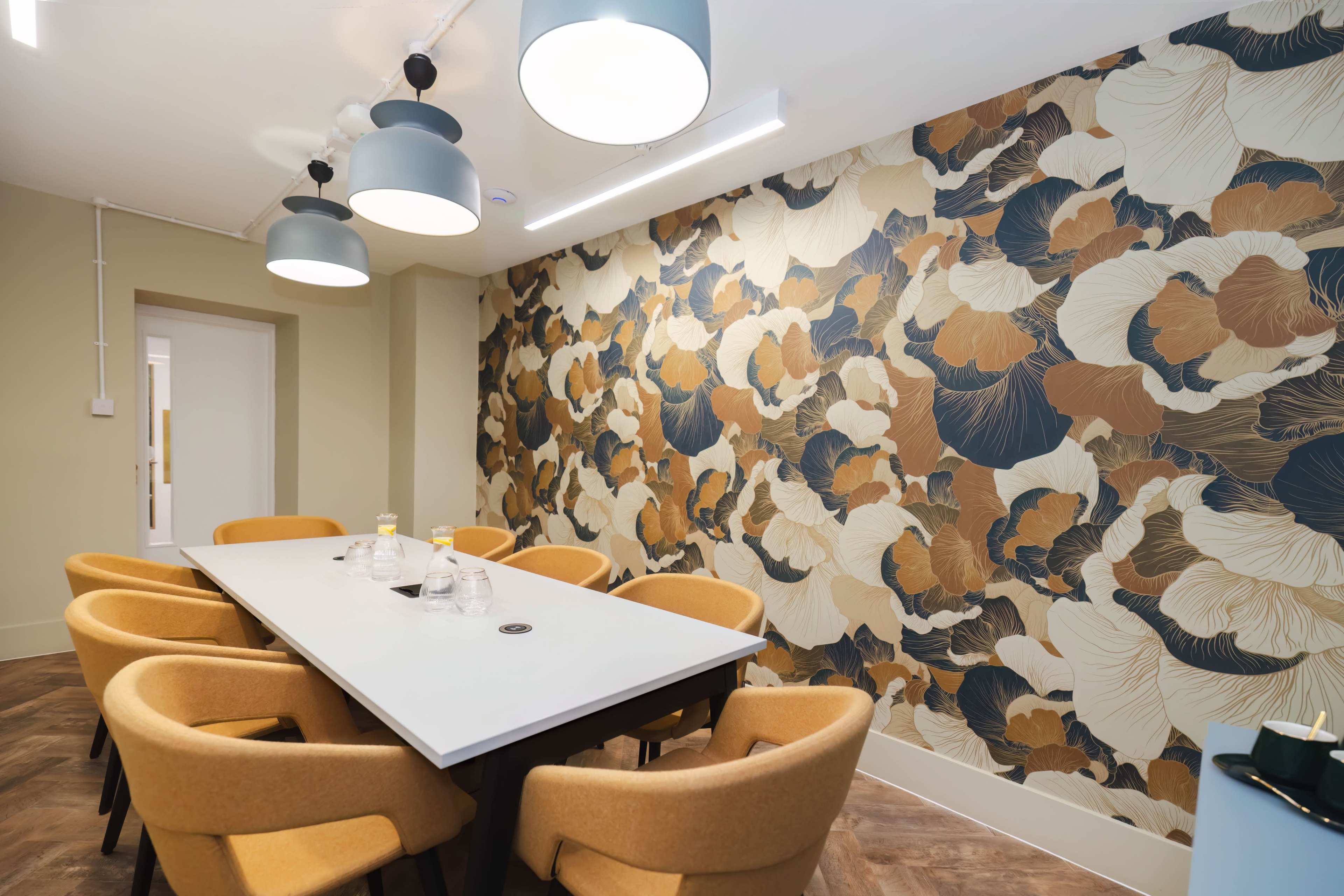 A modern conference room features a long table surrounded by yellow chairs, complemented by a patterned wallpaper and pendant lights.