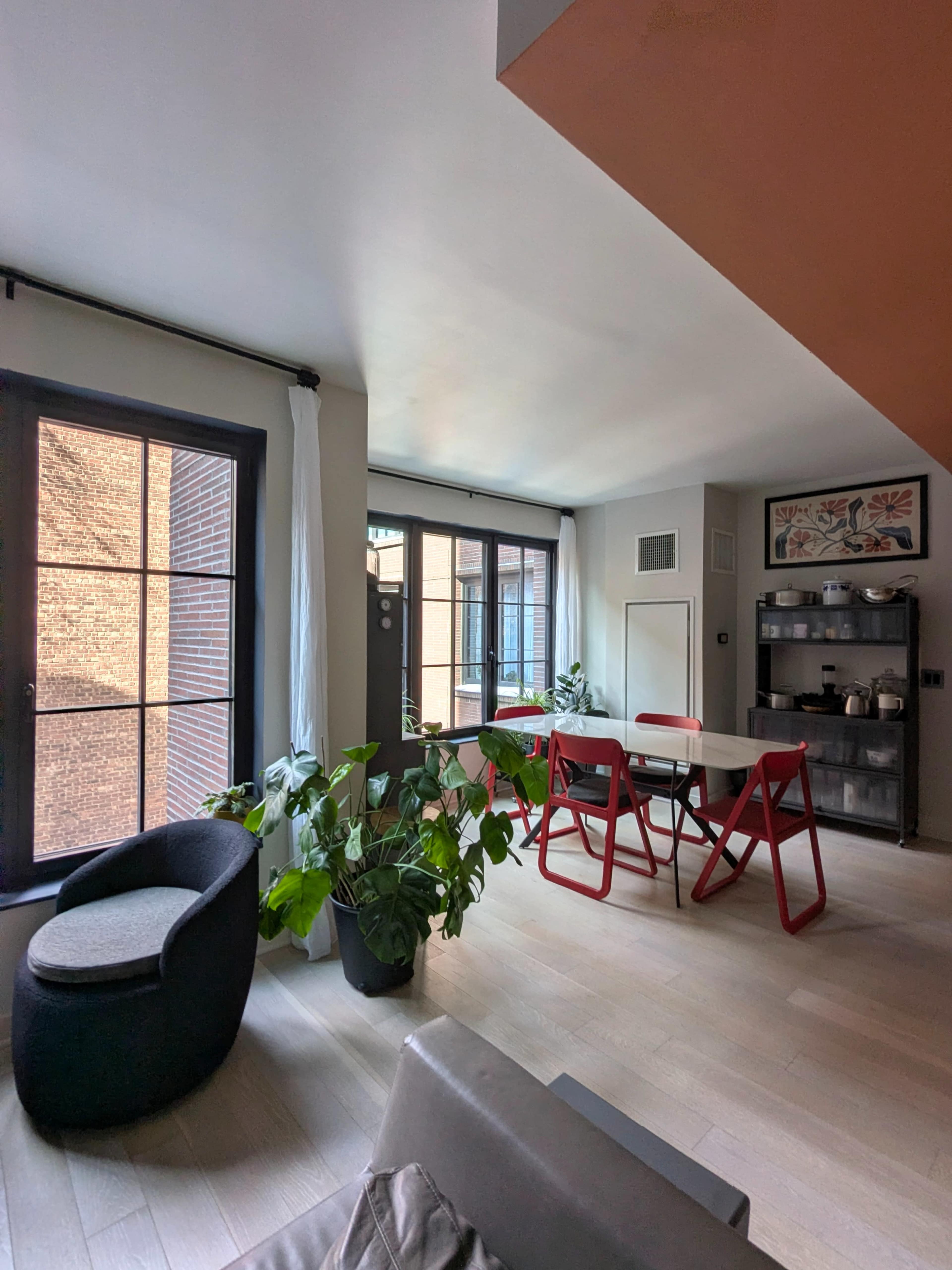 The image shows a modern living space featuring large windows, a dining area with a table and red chairs, and a black armchair next to a potted plant.