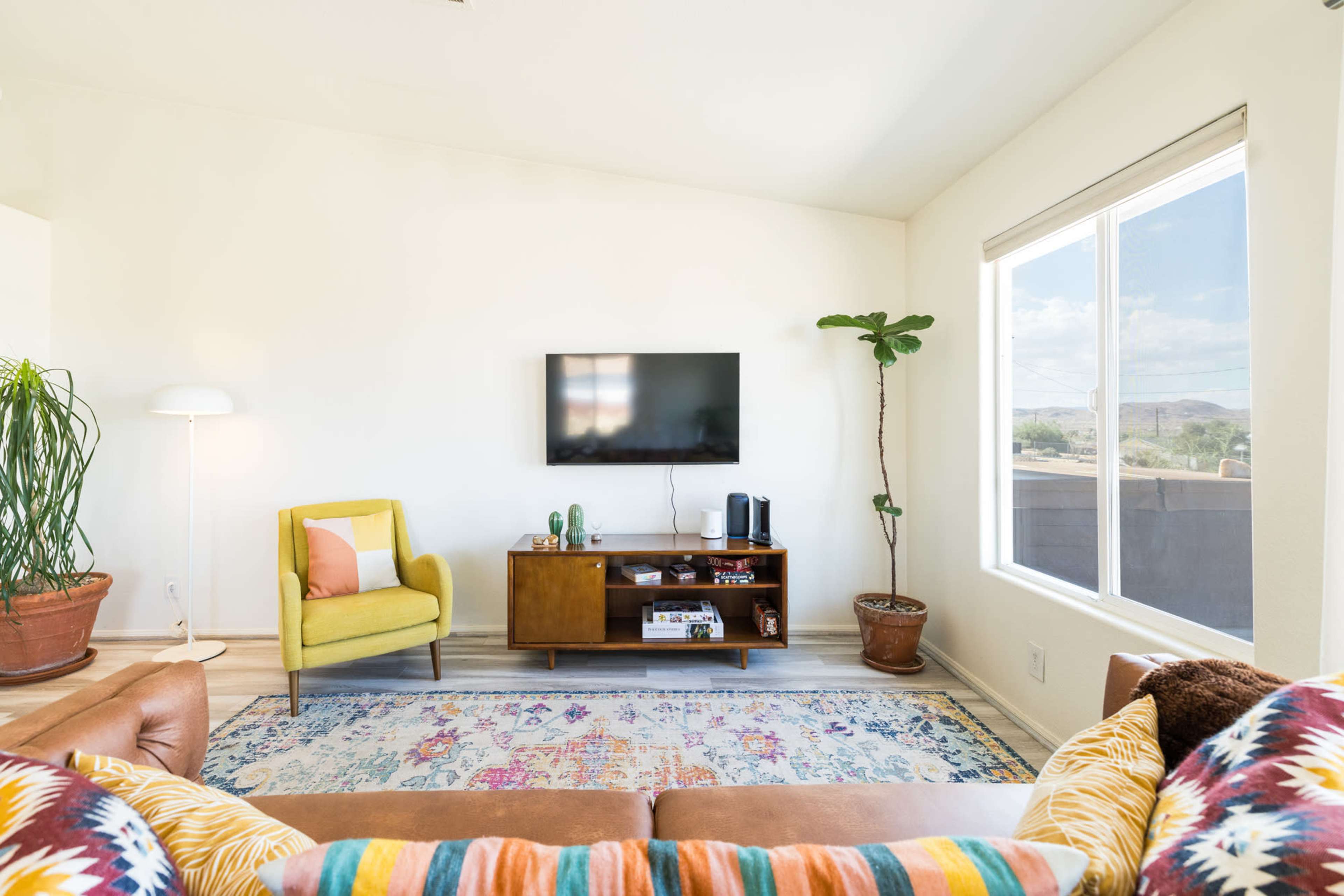 A bright living room features a yellow chair, a wooden TV stand with a television, and a large window providing natural light.