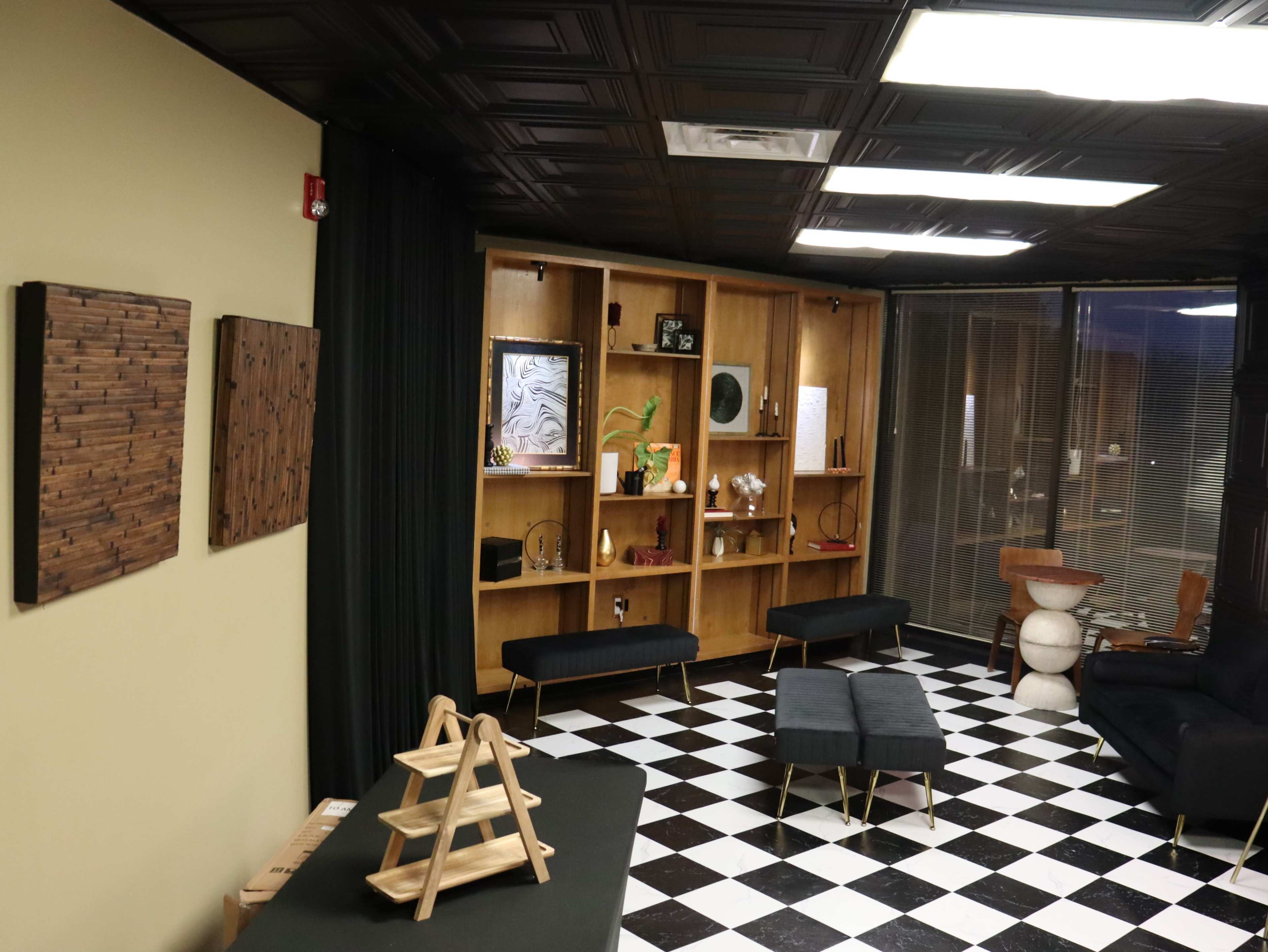 The image shows a room with a black and white checkered floor, featuring modern furniture, a wooden shelving unit with decorative items, and dark curtains.