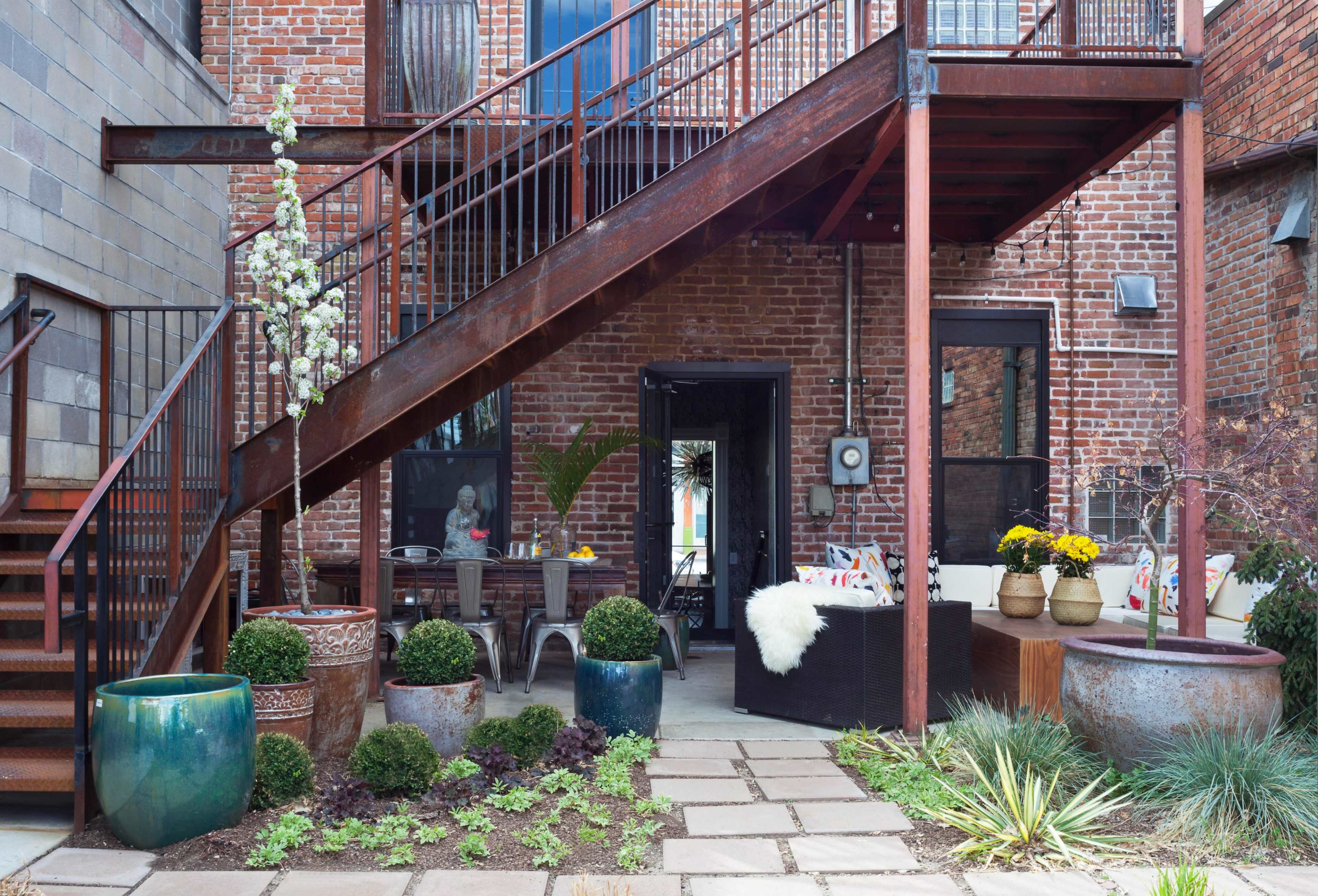 A metal staircase leads to a second-level patio area filled with potted plants, outdoor furniture, and a garden path made of stone tiles.