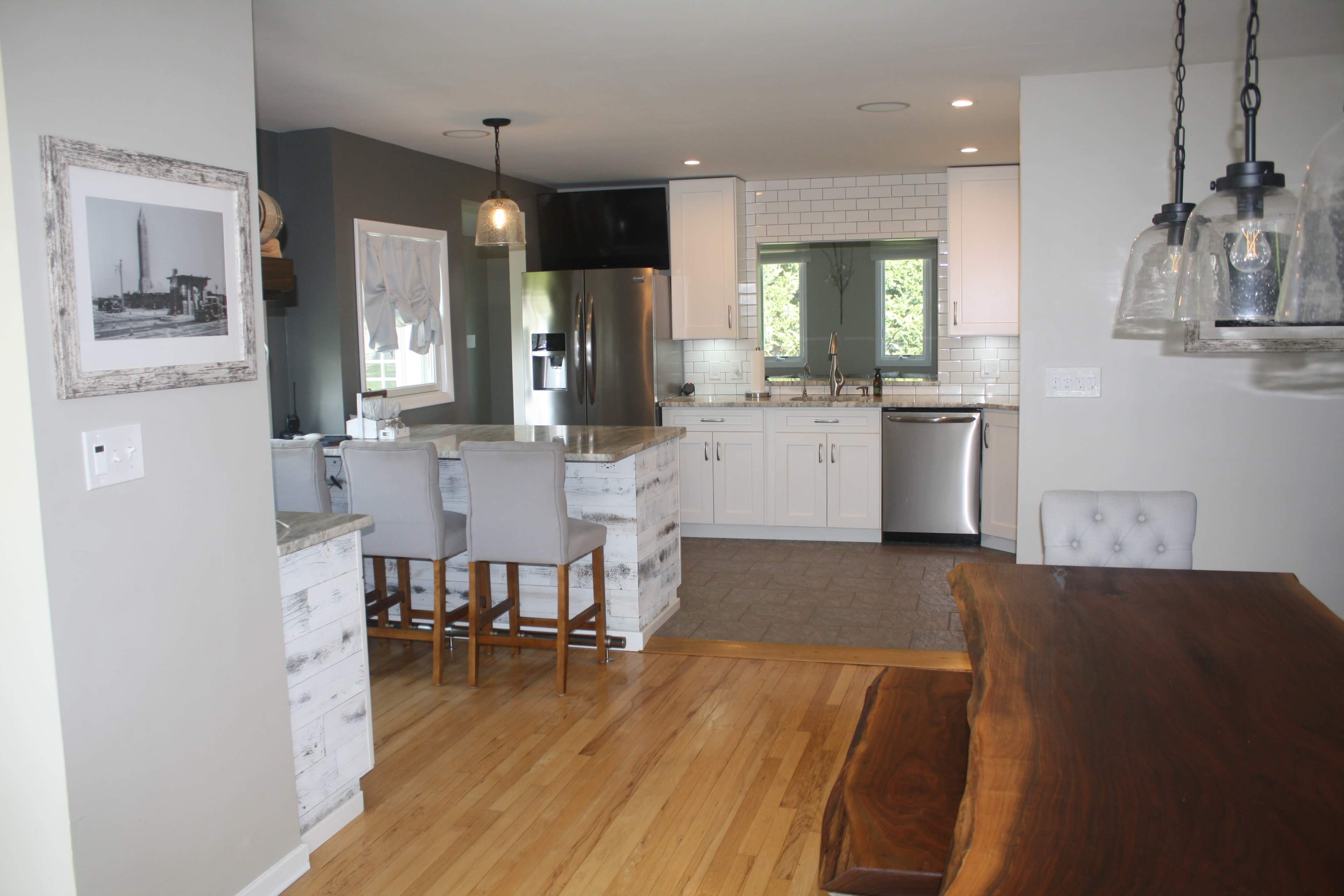 A modern kitchen area featuring white cabinets, a gray tile floor, a large wooden dining table, and an adjacent seating counter with bar stools.