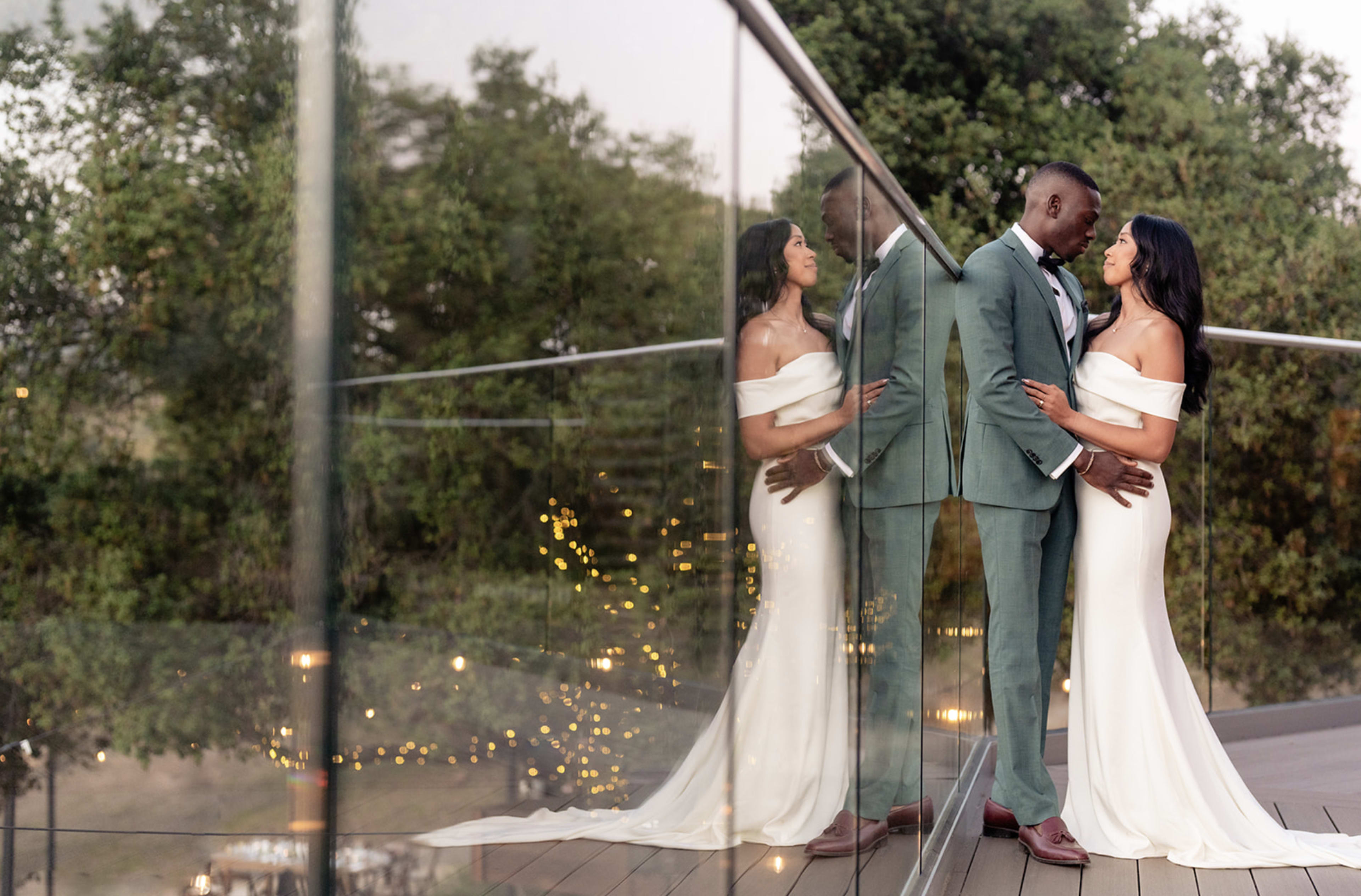 A couple stands close together on a balcony, with their reflections visible in the glass railing, surrounded by greenery and soft lights in the background.