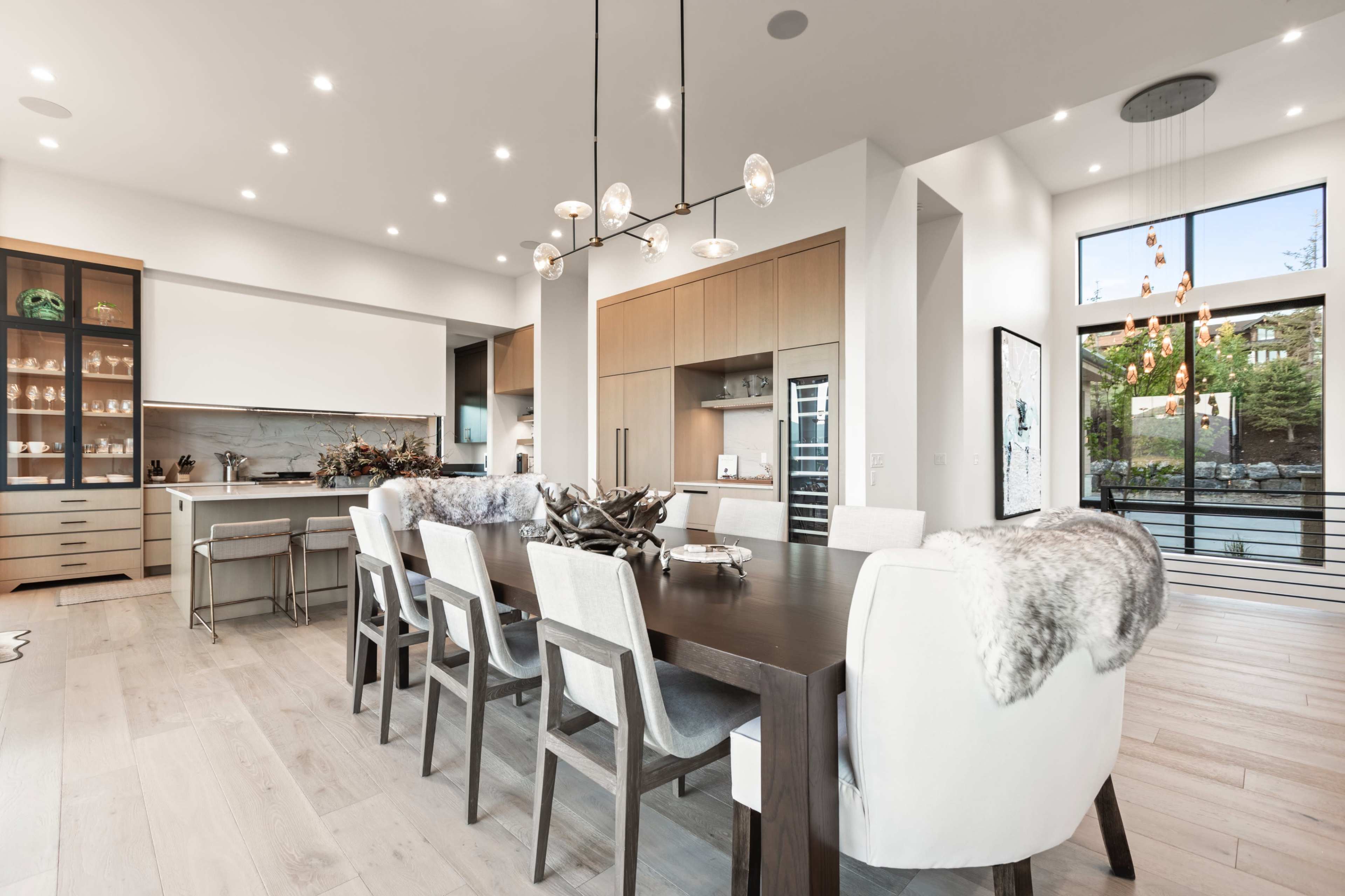 A modern dining area features a large wooden table surrounded by upholstered chairs, with a pendant light overhead and large windows letting in natural light.