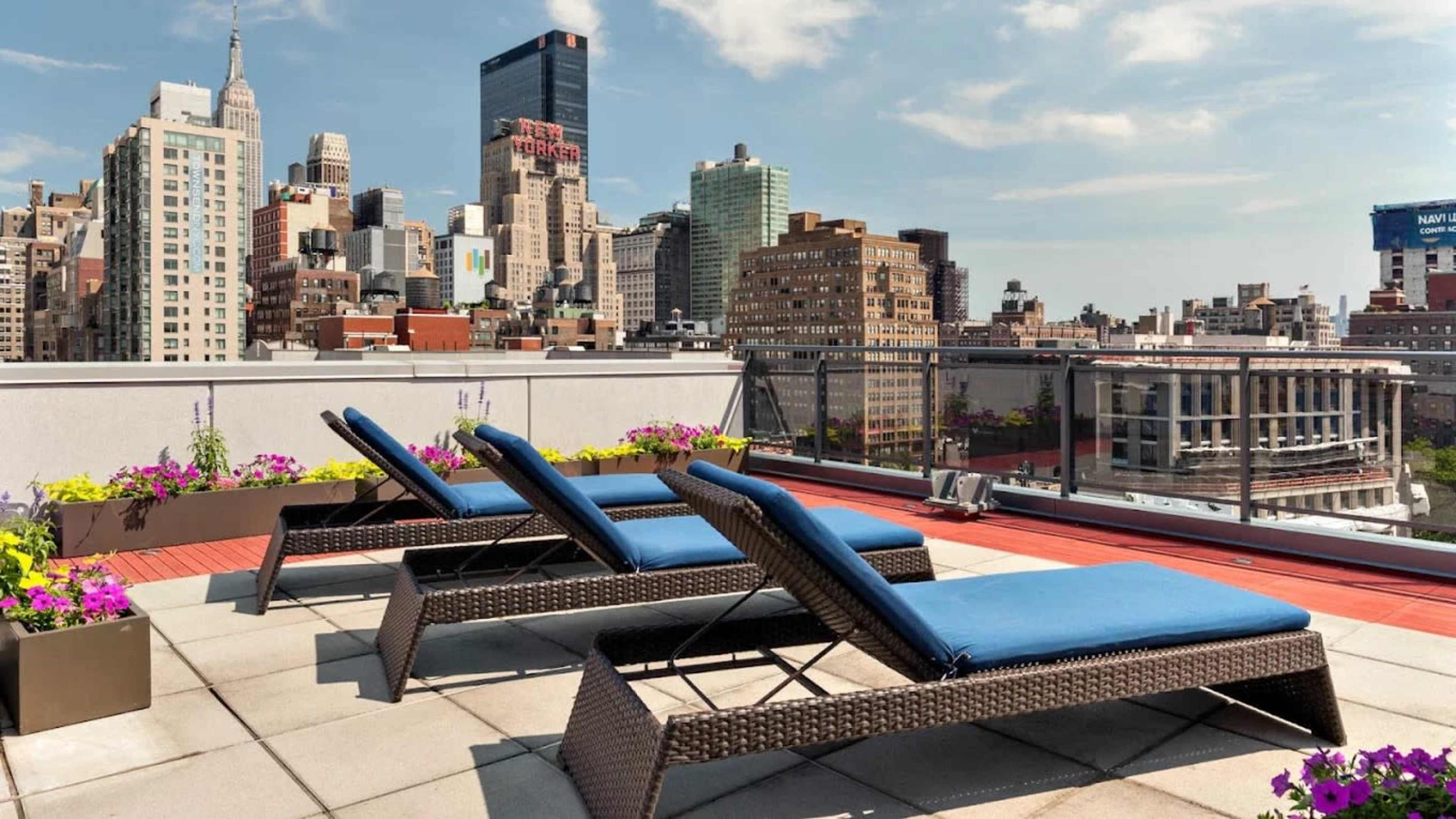 The image depicts a rooftop terrace with three lounge chairs overlooking a city skyline filled with various high-rise buildings.