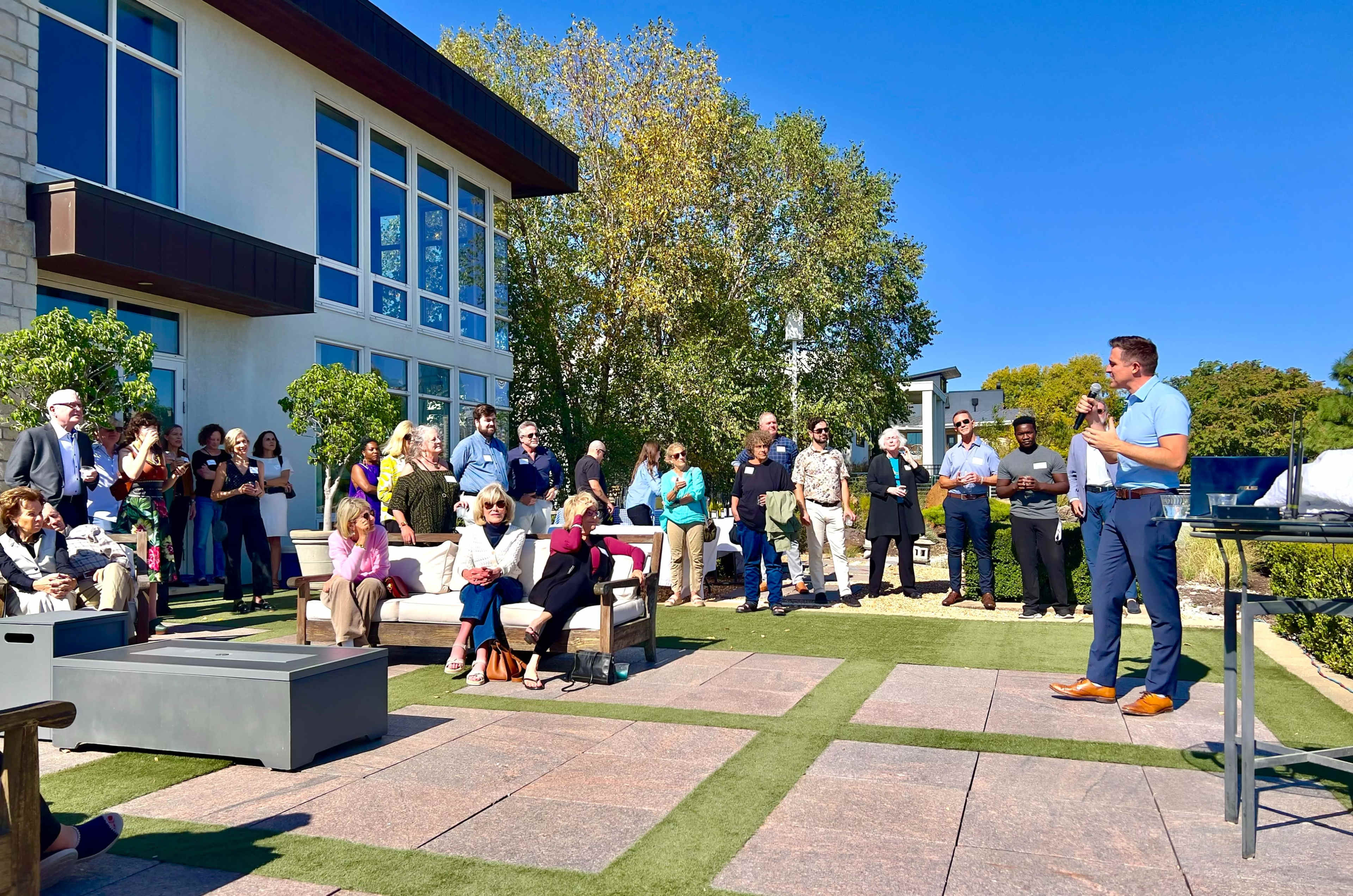 A speaker addresses a crowd gathered in an outdoor area with modern furniture and a clear blue sky.