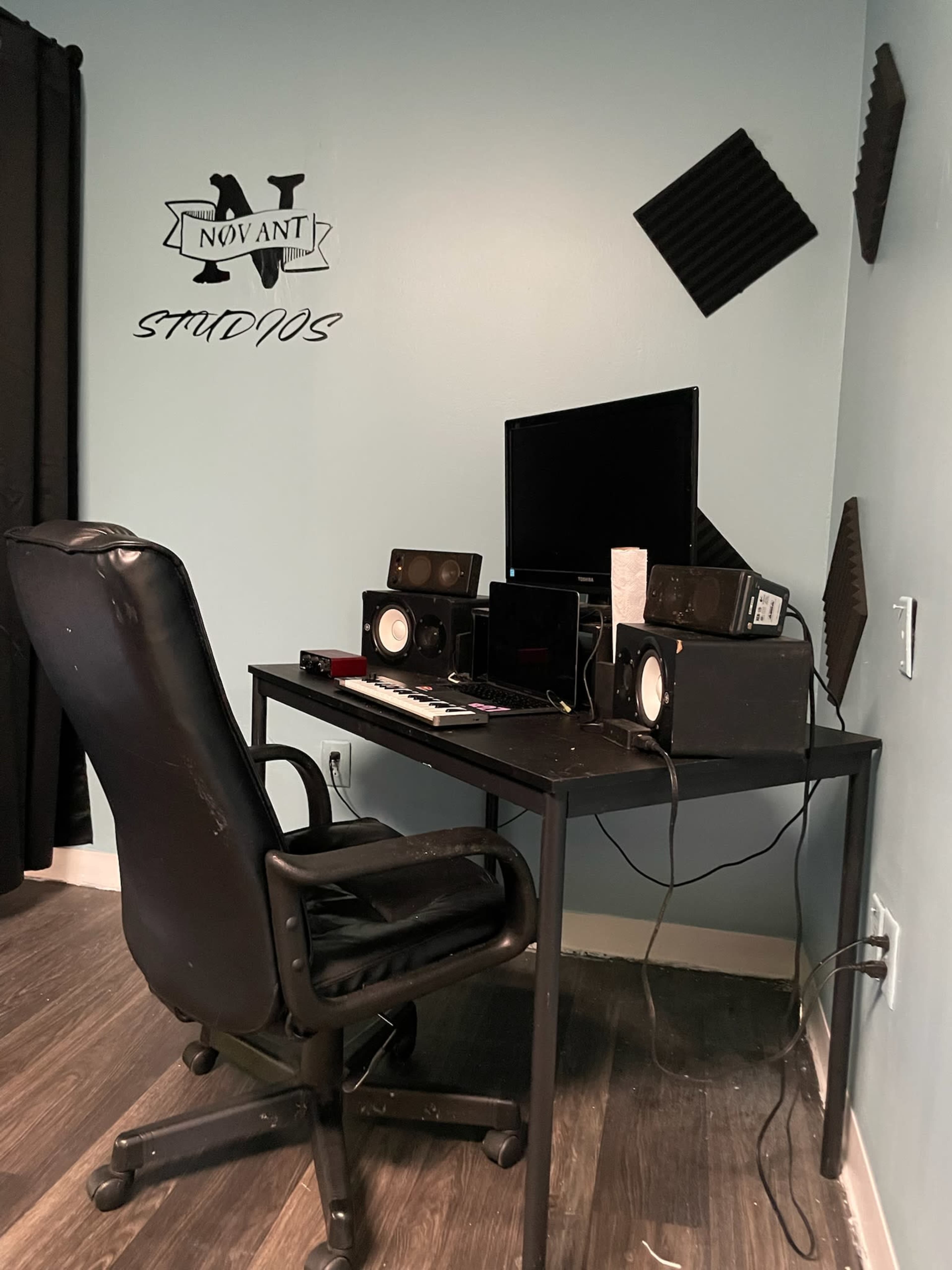 A black office chair sits in front of a desk with audio equipment and a monitor against a light blue wall with a decorative sign.