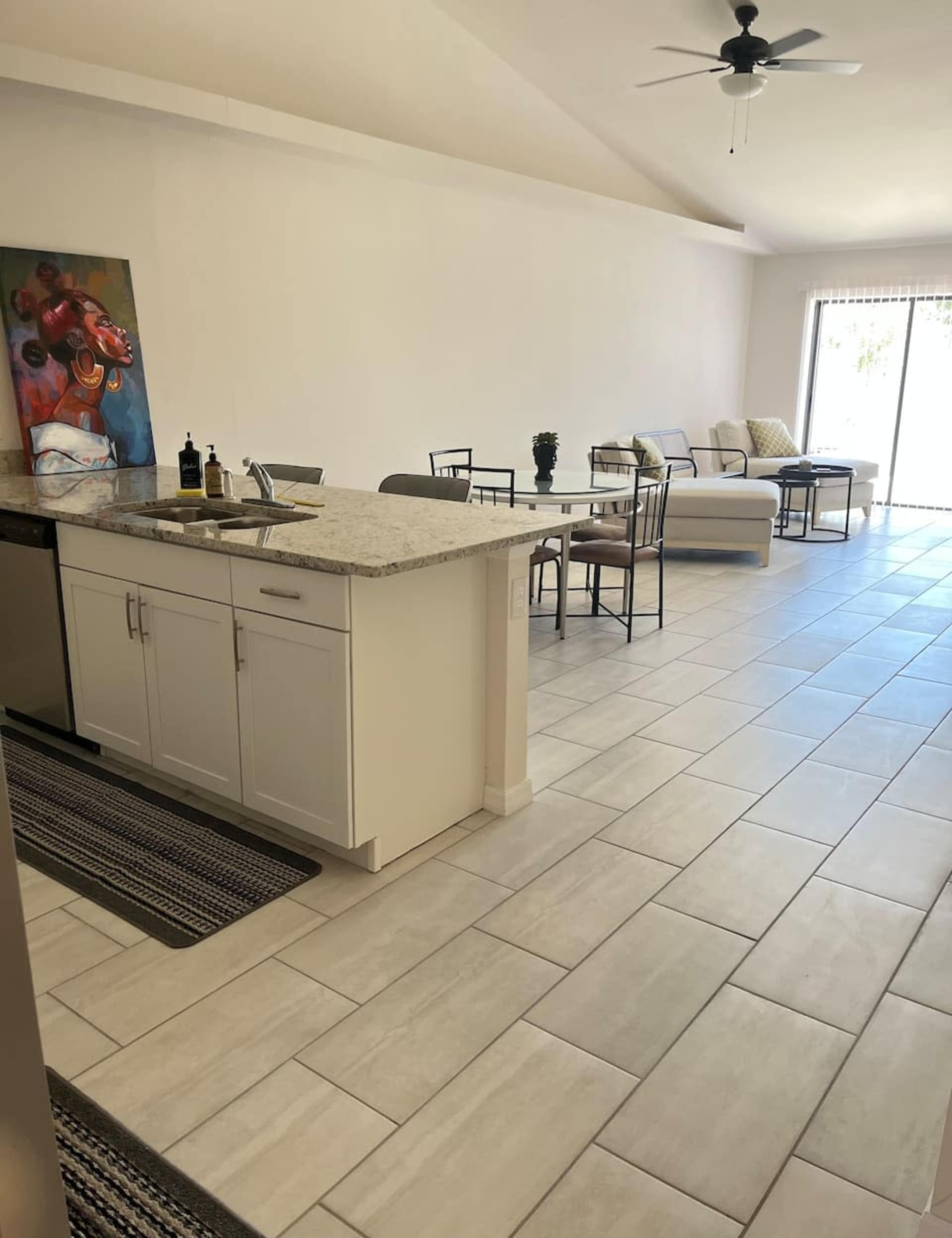 The image shows a modern kitchen area with a granite countertop, dining table, and a living space featuring a sofa and large sliding glass door to the outside.