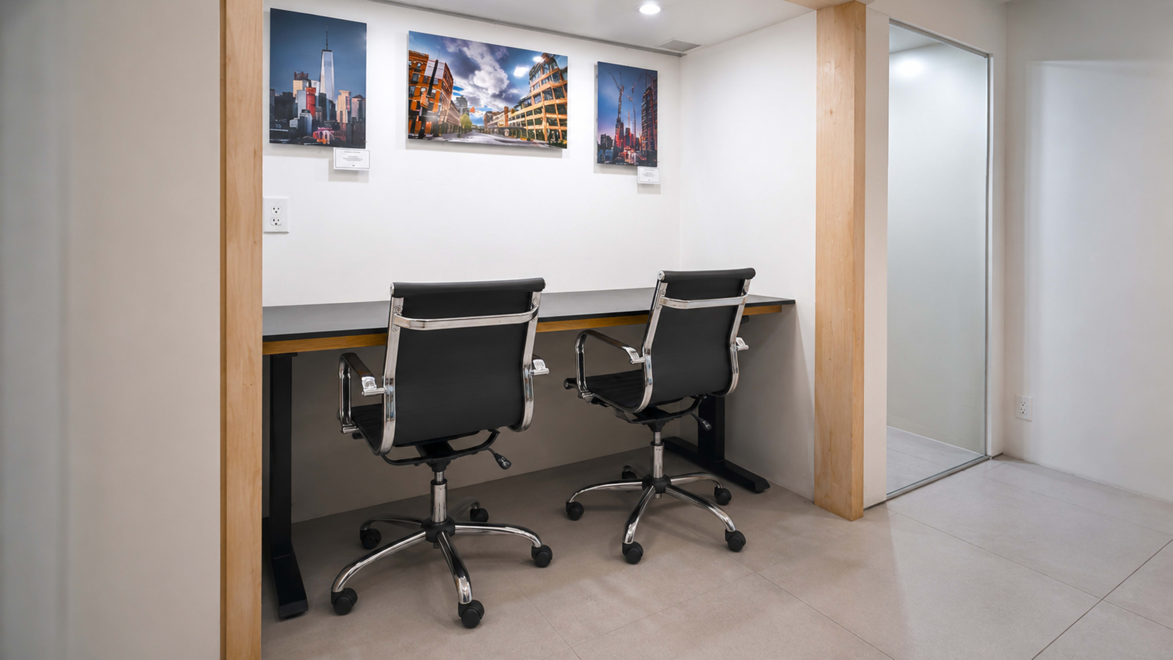 The image shows a modern workspace with two black office chairs positioned at a shared desk against a wall featuring three framed photographs.