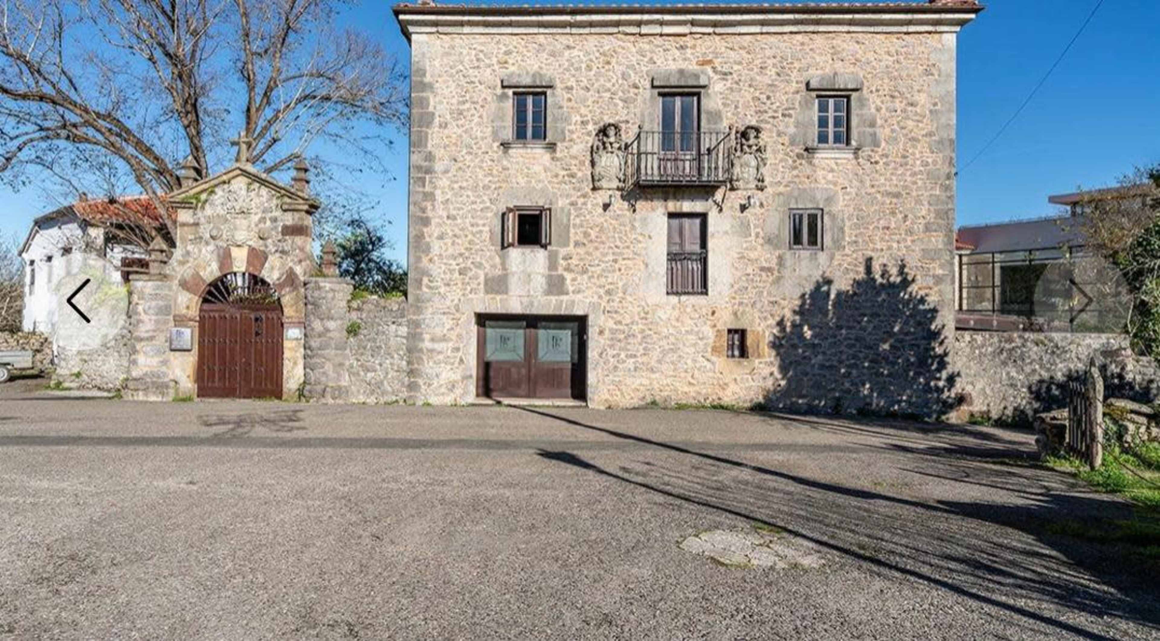 The image shows a stone building with a symmetrical facade, two balconies, and an entrance marked by a wrought iron gate, set in a rural landscape with clear blue skies.