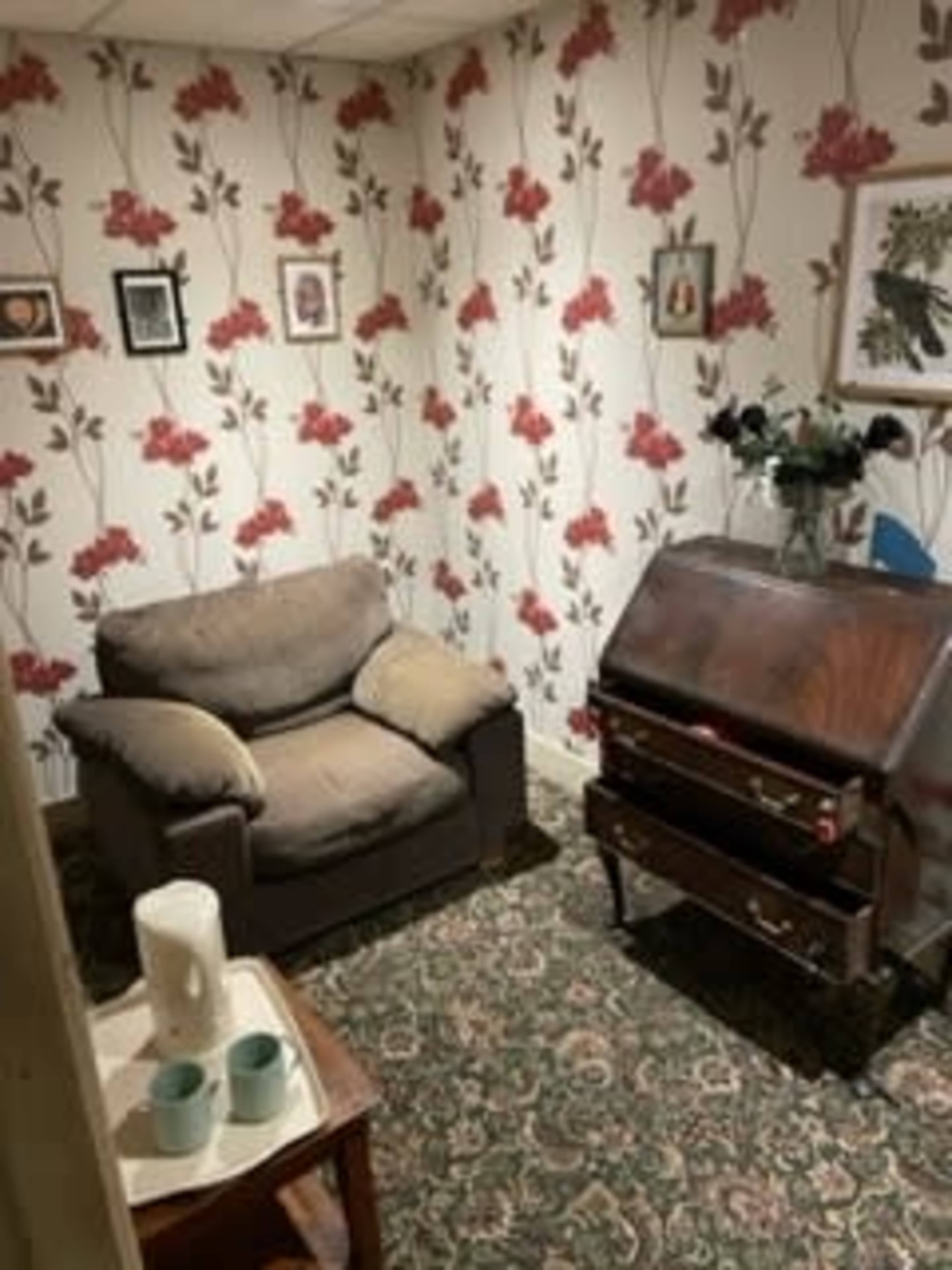 A small room with floral wallpaper, a brown sofa with cushions, a wooden desk, and a small table with cups and a white vase.