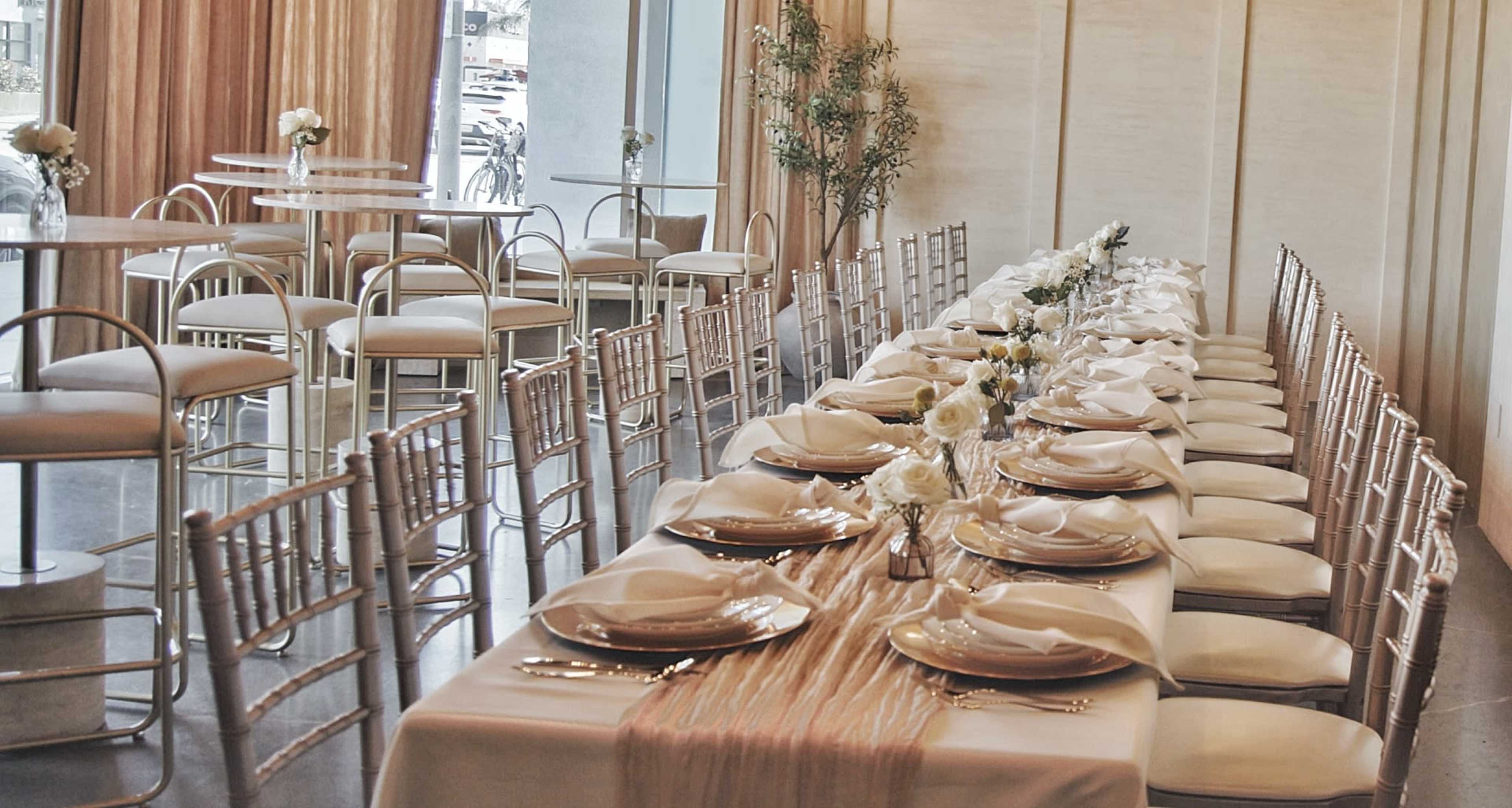 A long dining table is elegantly set with white tableware and gold accents, surrounded by rows of wooden chairs in a bright, modern room.