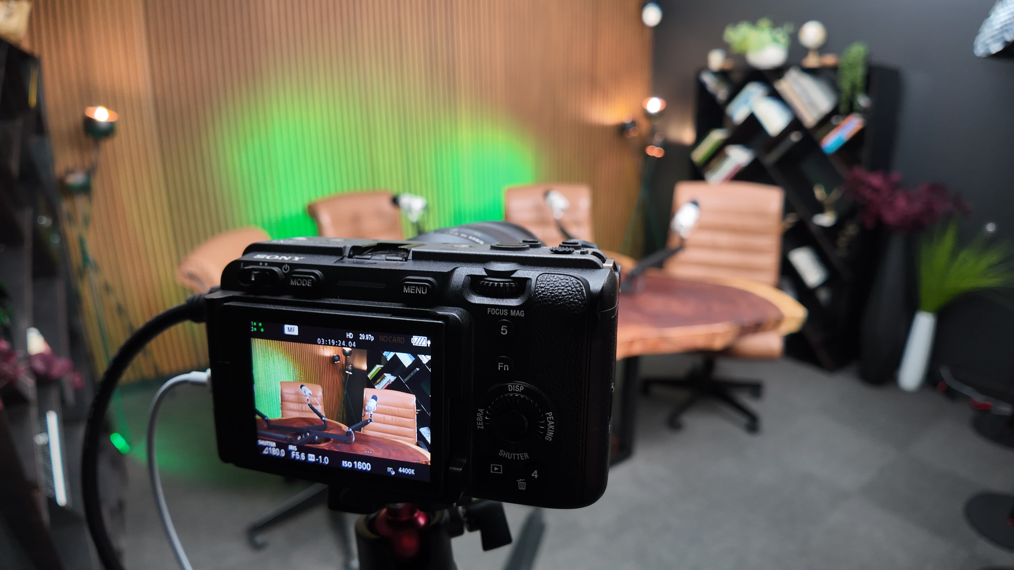 A camera is positioned on a tripod, capturing a well-lit studio setup featuring two brown chairs, a round table, and a backdrop with decorative elements.