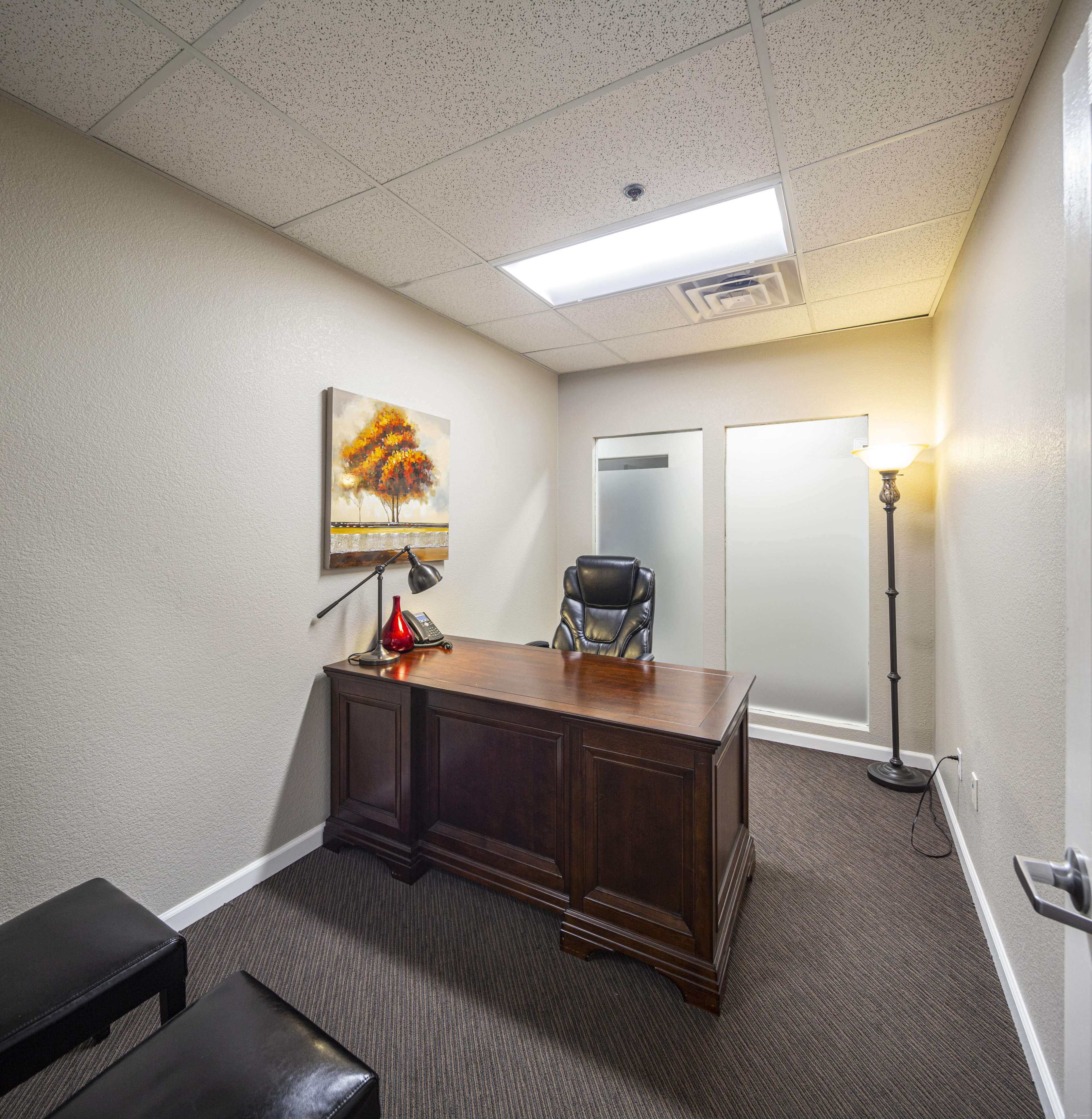 A small office features a dark wooden desk with a chair, a painting on the wall, and a floor lamp in the corner.