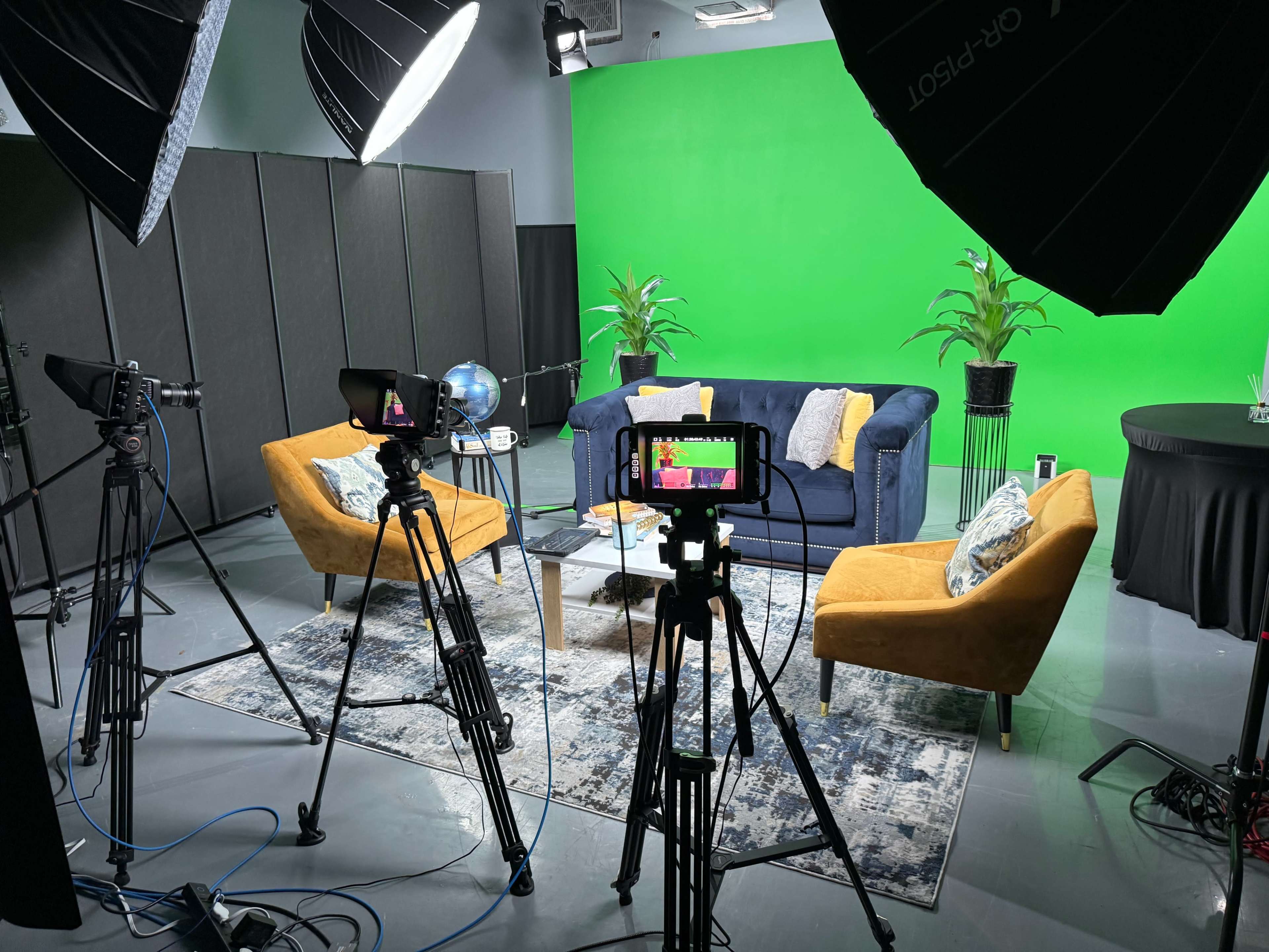 The image shows a television studio setup featuring two cameras pointed at a modern seating area with a blue couch and two yellow chairs, surrounded by lighting equipment and green backdrop.